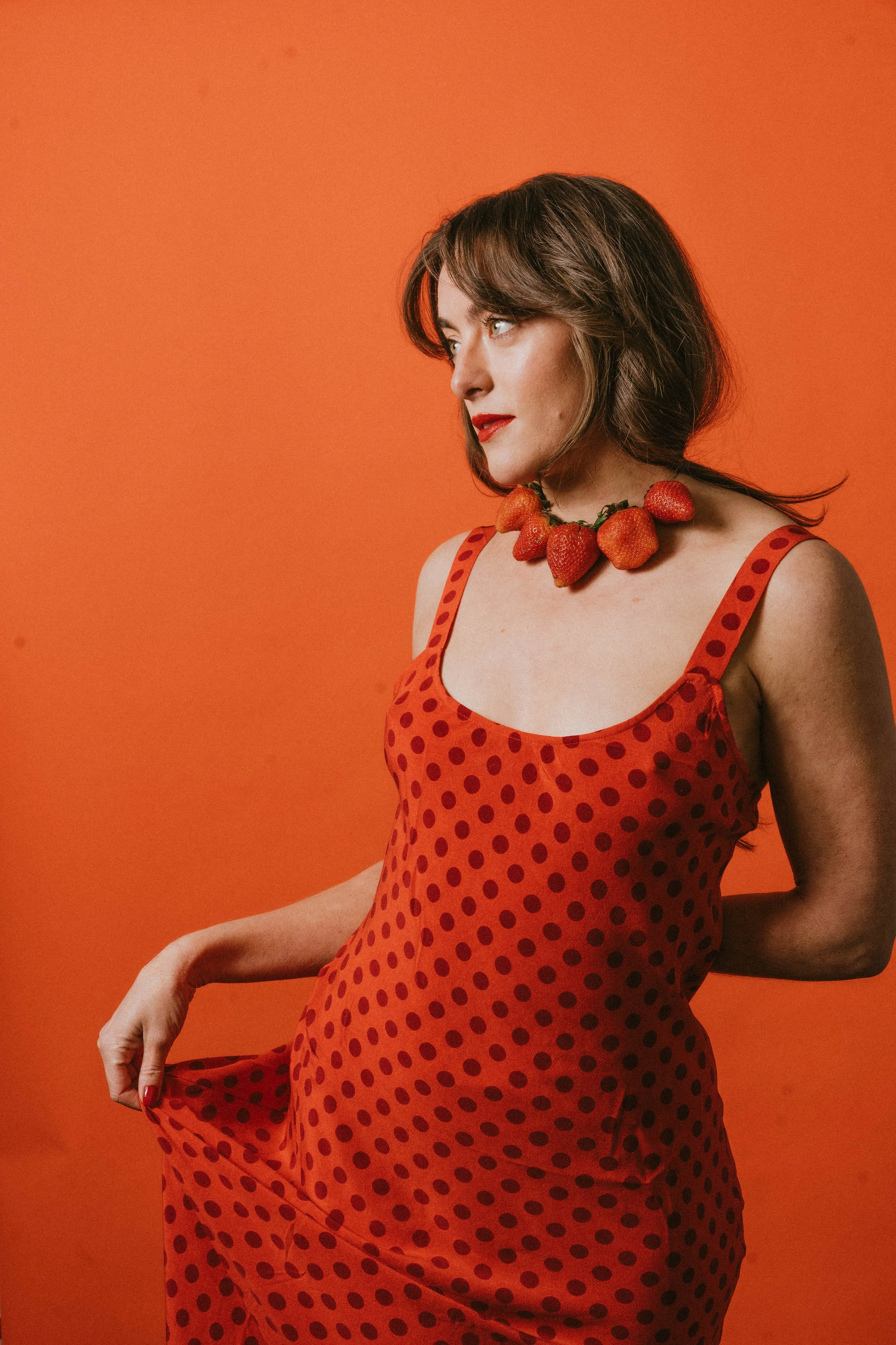 A woman in a red polka dot dress holding the hem of her dress, wearing a necklace made of strawberries, against an orange background.