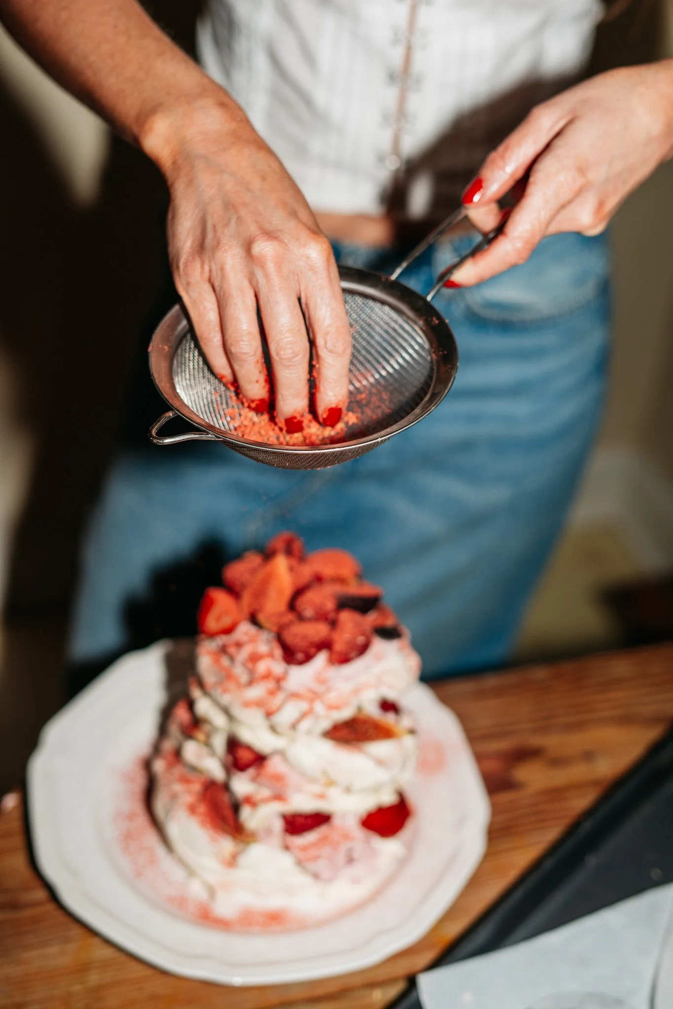 A person sprinkling pink-colored sugar or crushed candy over a strawberry shortcake dessert with whipped cream and strawberries, placed on a white platter on a wooden surface.