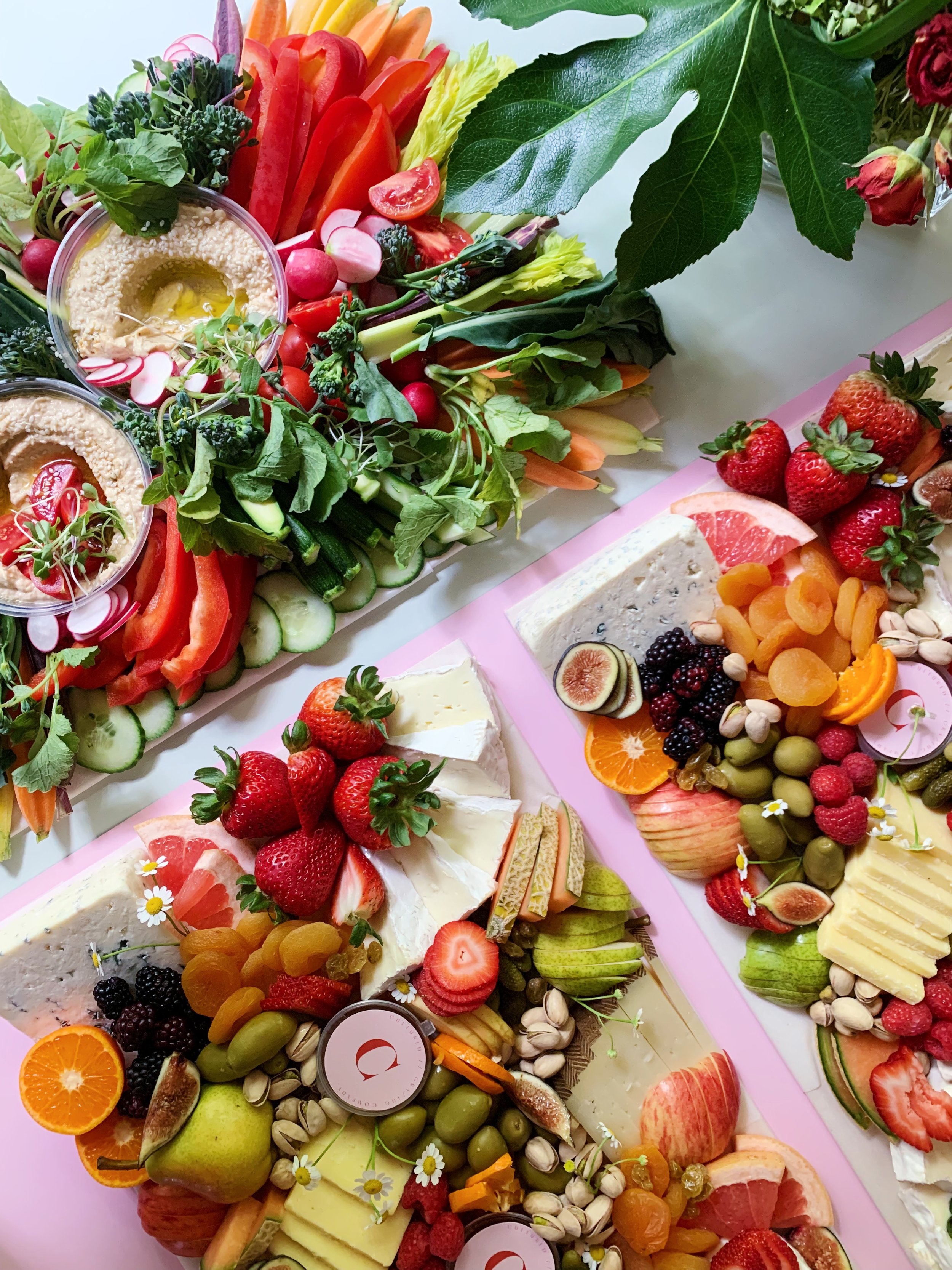Assorted fruit and vegetable platters with strawberries, grapes, oranges, apples, tomatoes, radishes, cucumbers, carrots, and leafy greens, along with cheese, hummus, and garnishes.