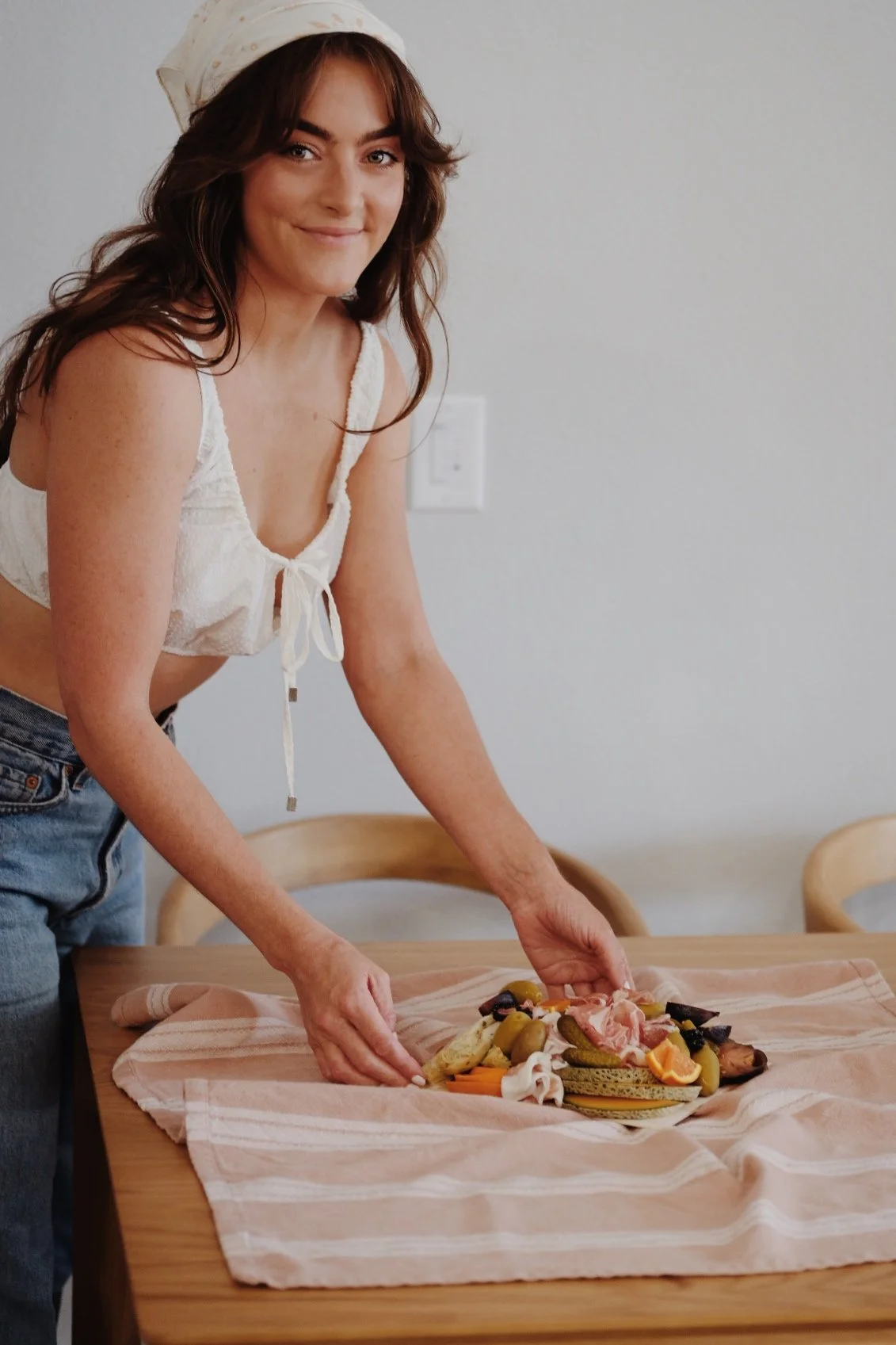A young woman with brown hair, wearing a white top and a white headscarf, is preparing a charcuterie board on a wooden table.