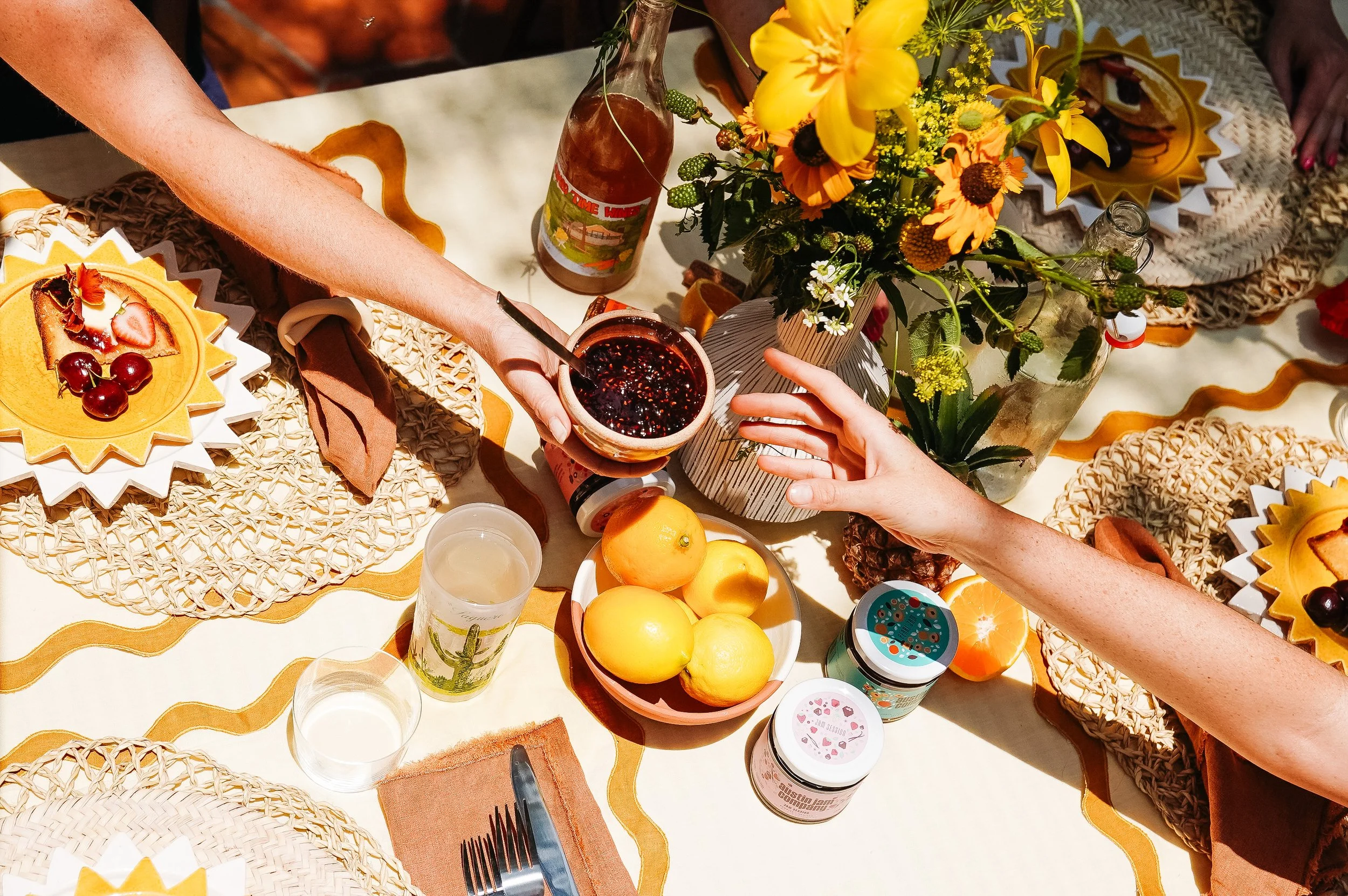 Top-down view of a table set for a meal, with a bouquet of yellow flowers, plates of sliced fruit, jars, glasses, and hands reaching for food.