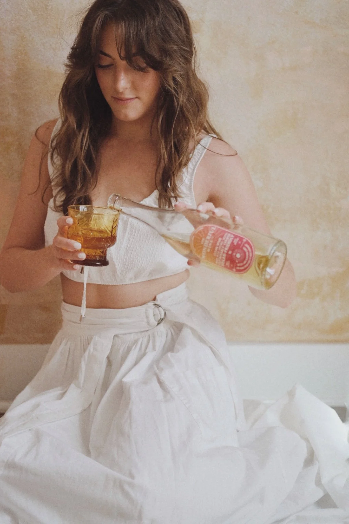 Woman pouring a beverage from a bottle into a glass, wearing a white crop top and white skirt, against a beige background.