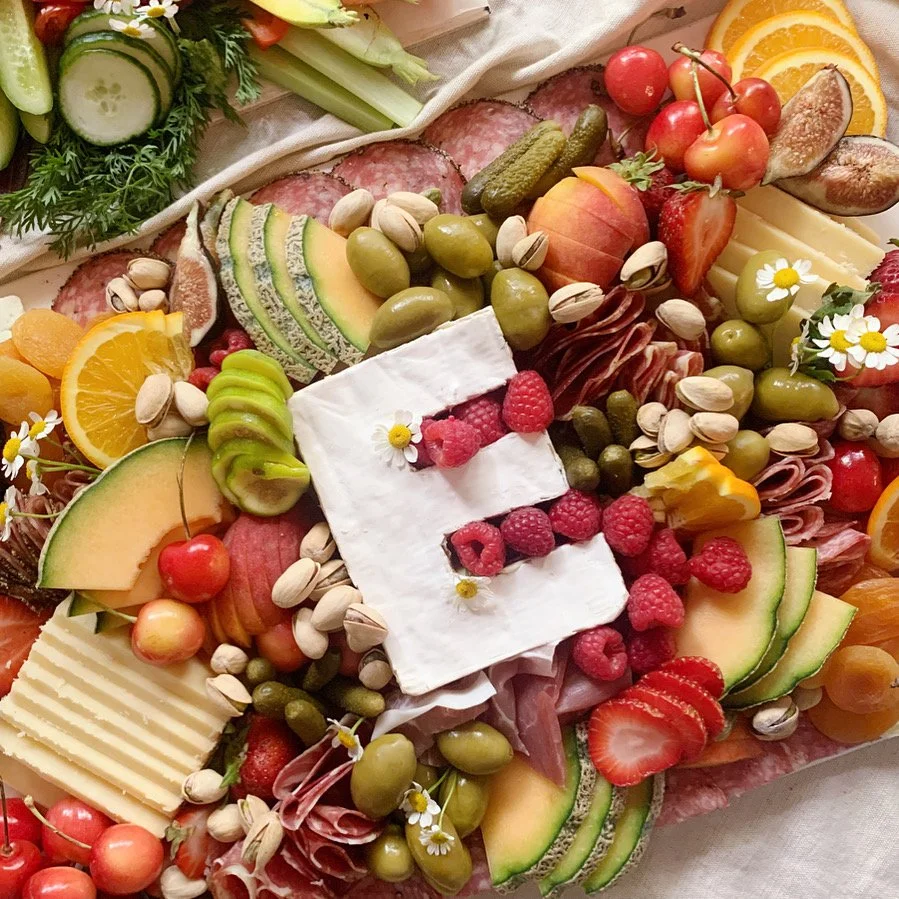 Charcuterie board with assorted cheeses, fruits like strawberries, raspberries, cherries, orange slices, melon, grapes, and figs, along with nuts, pickles, and flowers.