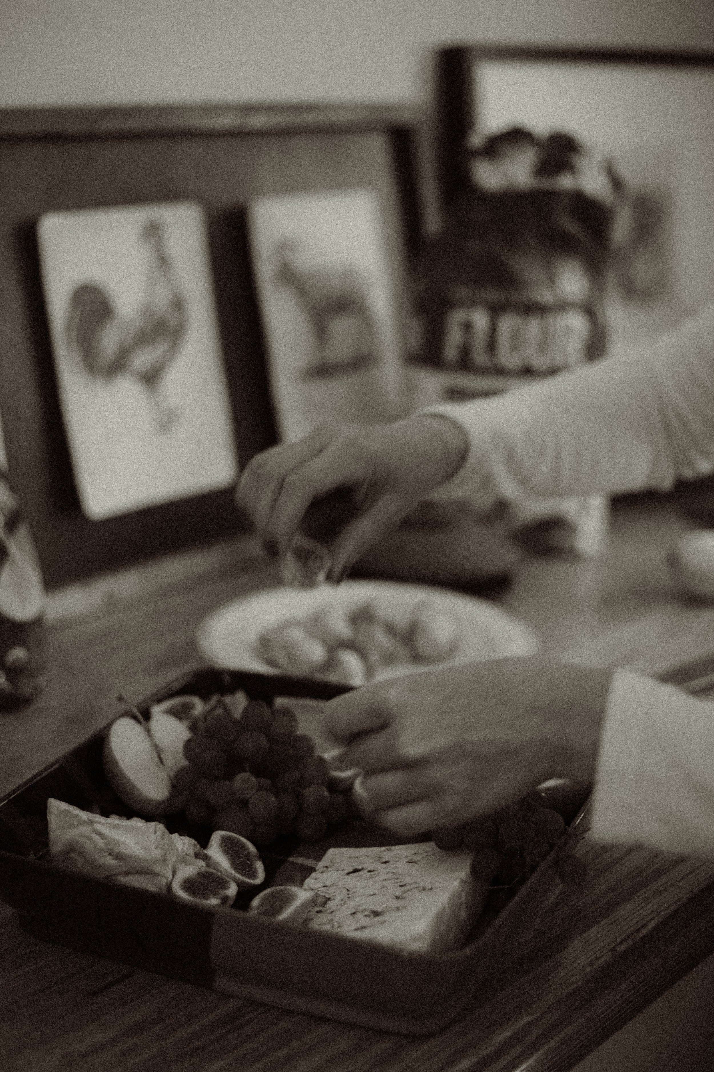 Person reaching for food on a tray that includes grapes, figs, and cheese, with framed animal pictures and a flower pot in the background.