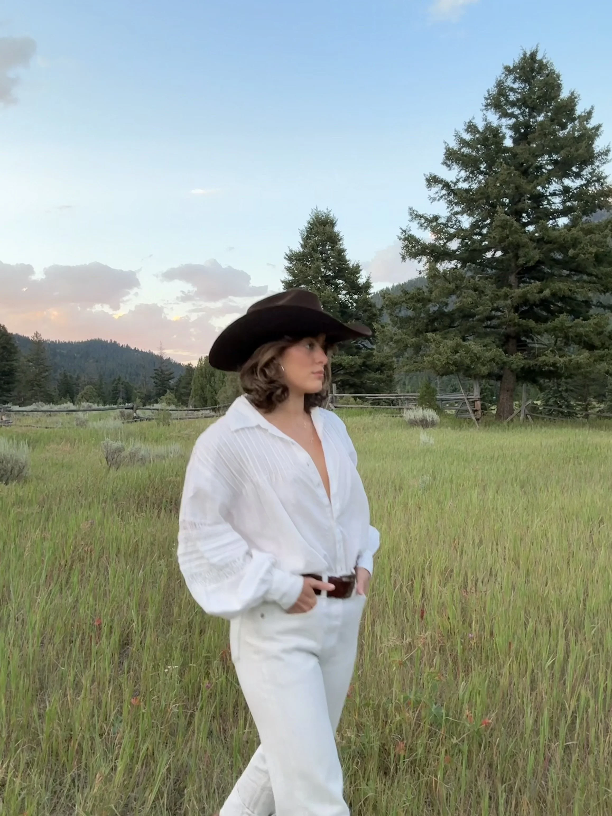 A woman in a white outfit and wide-brimmed hat standing in a grassy field with trees and mountains in the background during sunset.
