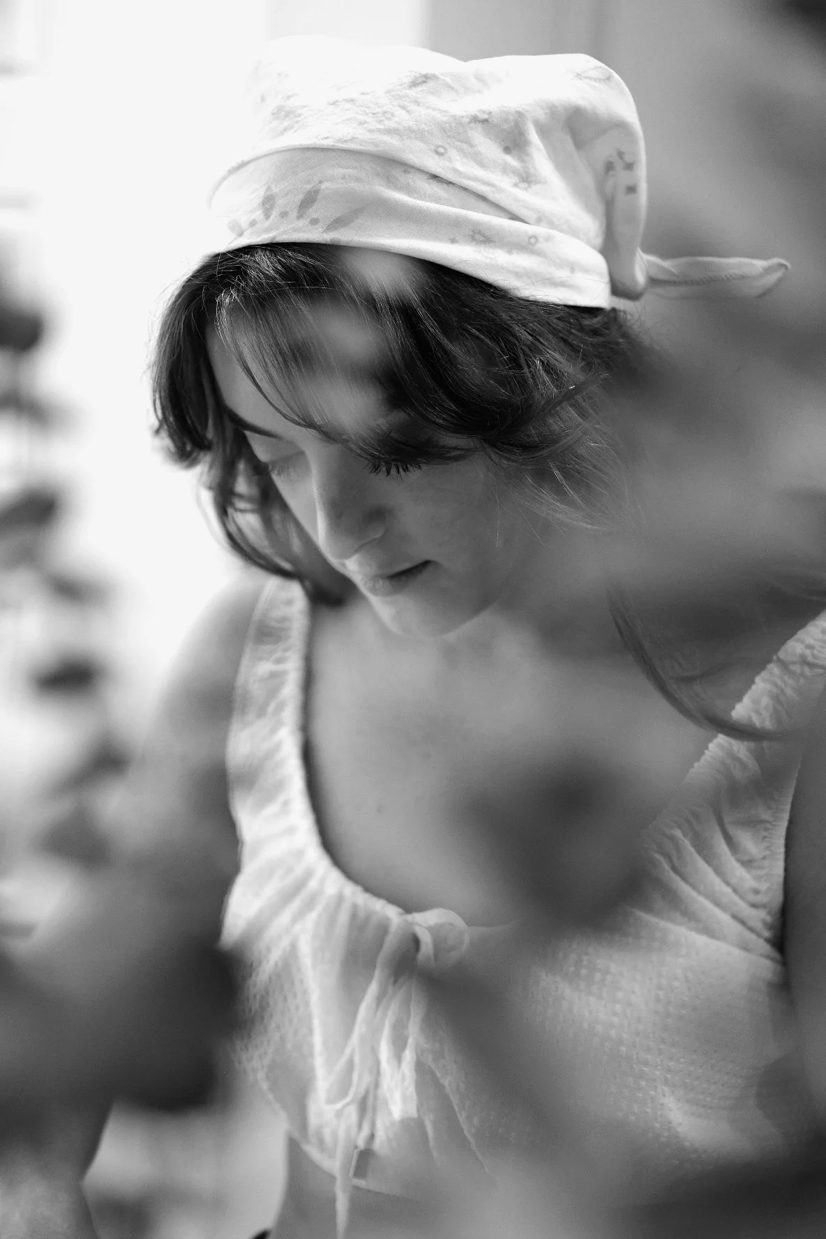 A woman with dark, wavy hair wearing a white headscarf and a sleeveless, textured top, looking downward with a soft expression.