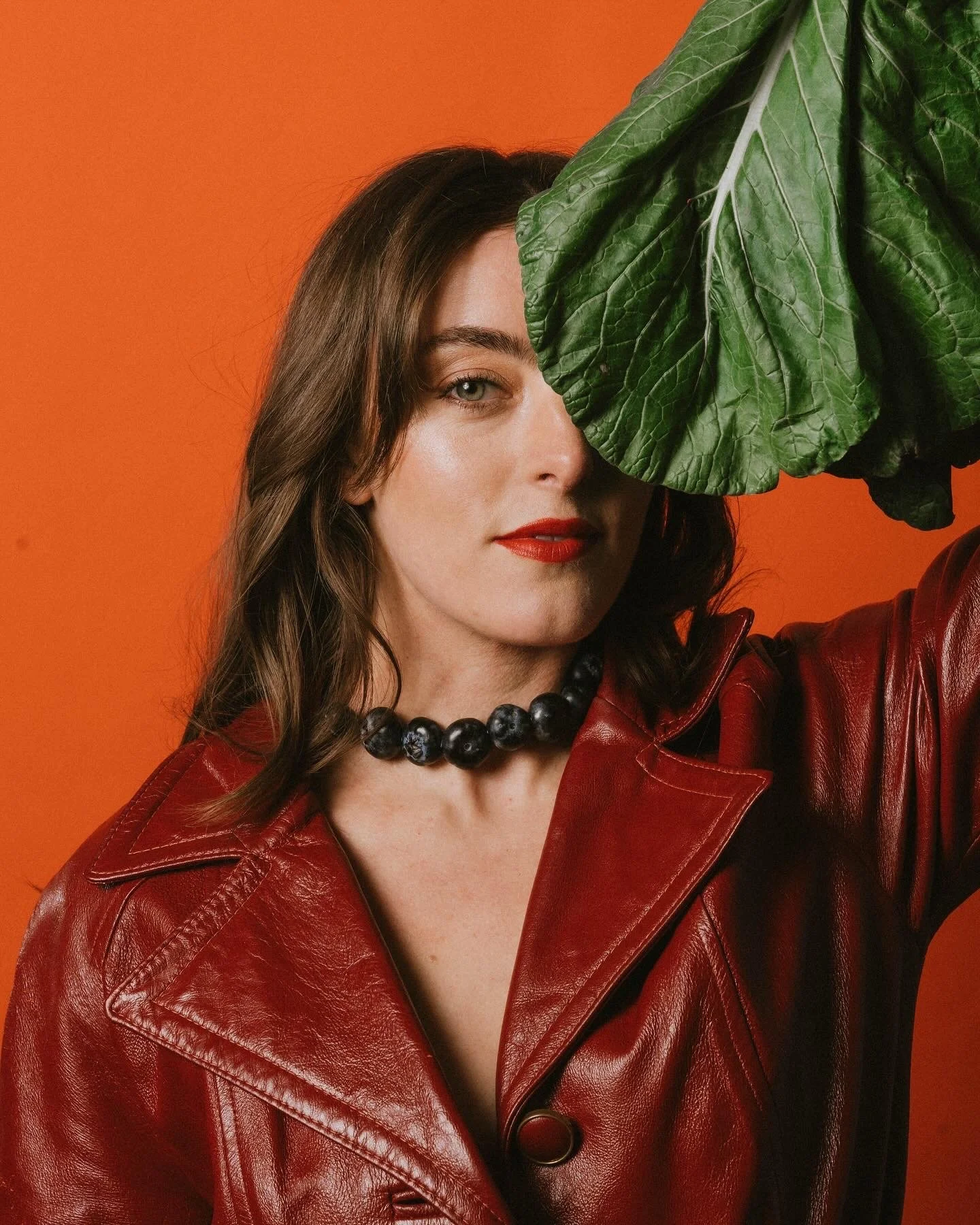 A woman with brown hair, wearing a red leather jacket and a black beaded necklace, is partially hidden by a large green leaf against an orange background.