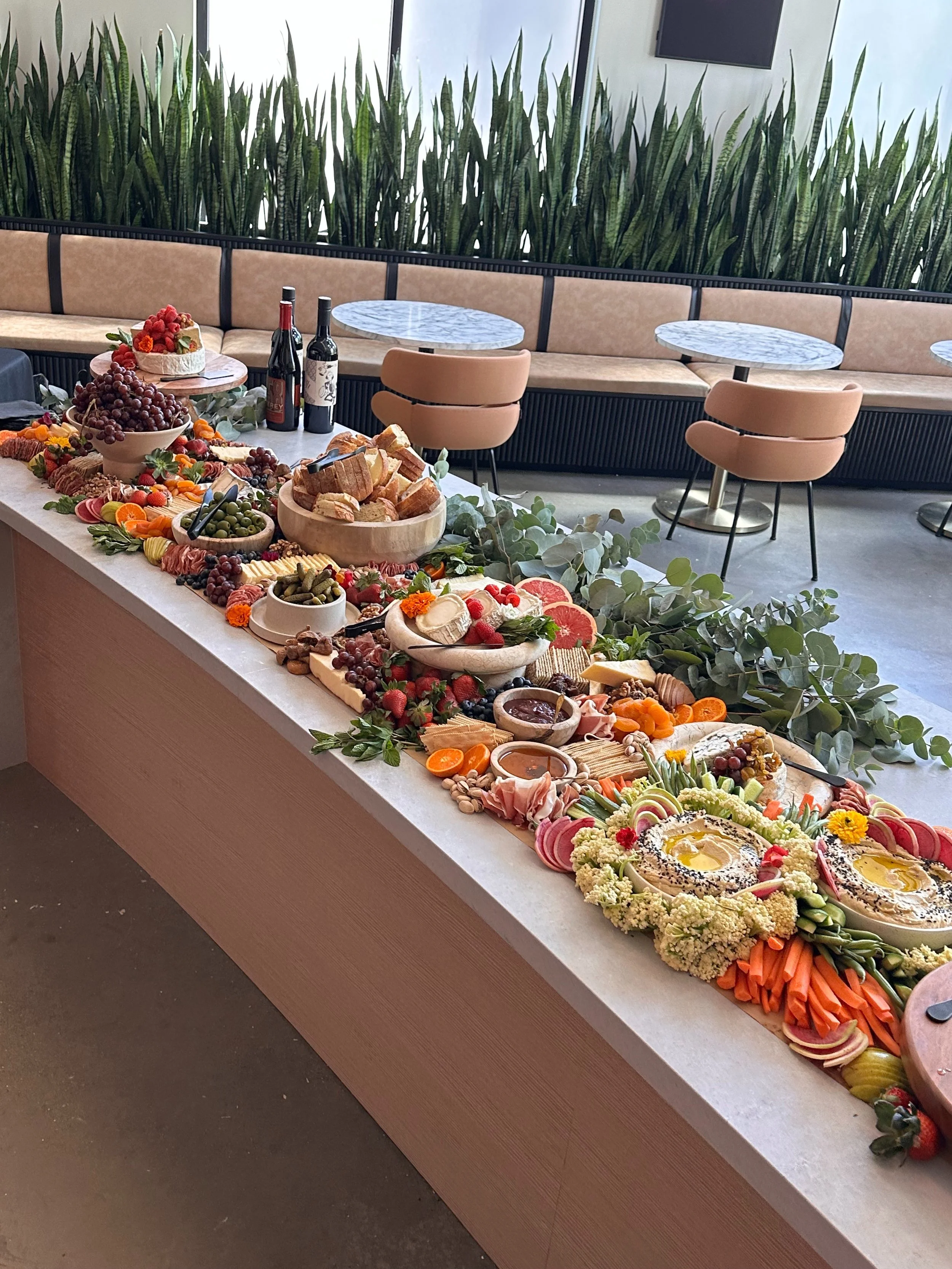 A long buffet table with various cheeses, fruits, vegetables, bread, jams, and wines set in a modern dining area with green plants, marble tables, and beige seating.