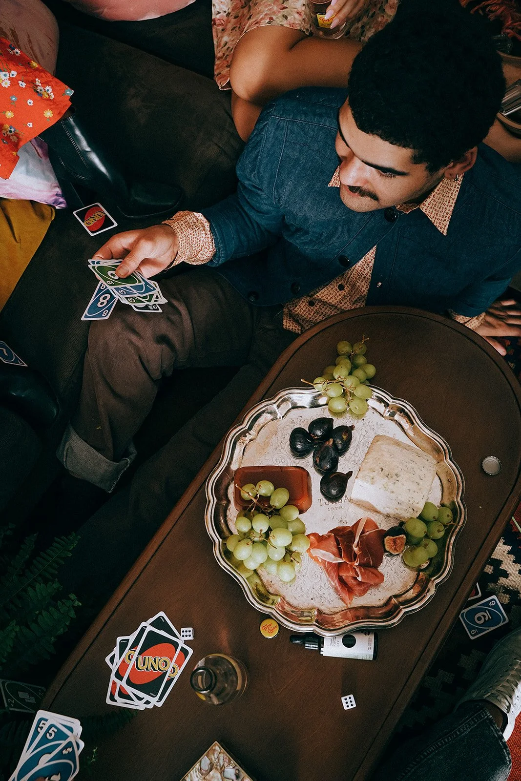 People playing card games around a table with snacks, grapes, cheese, and candy on it.