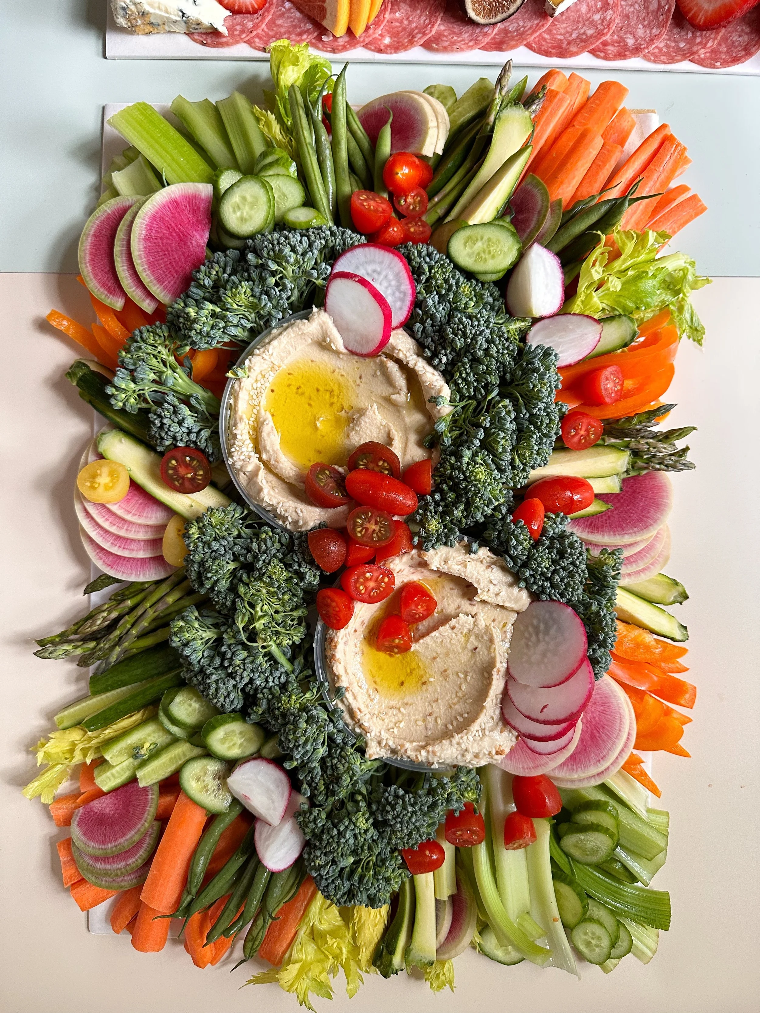 A platter of fresh vegetables including sliced radishes, cherry tomatoes, cucumbers, carrots, celery, and broccoli, with a bowl of hummus topped with olive oil and cherry tomatoes in the center.