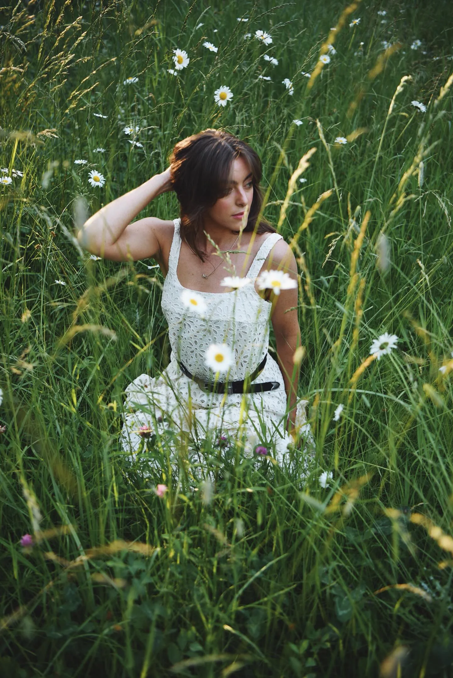 A young woman with brown hair sitting in a field of tall green grass and wildflowers, wearing a white eyelet dress.