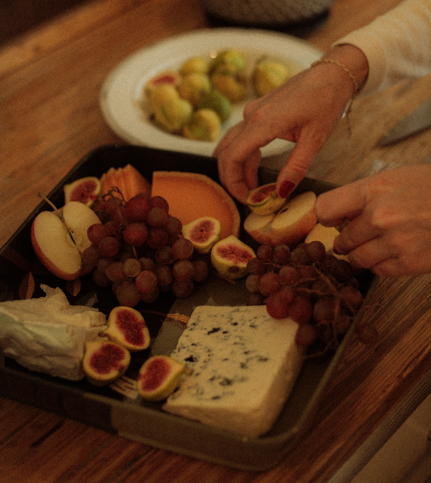 Hands arranging sliced figs, grapes, and cheese on a tray with a plate of grapes and a wedge of cheese nearby.