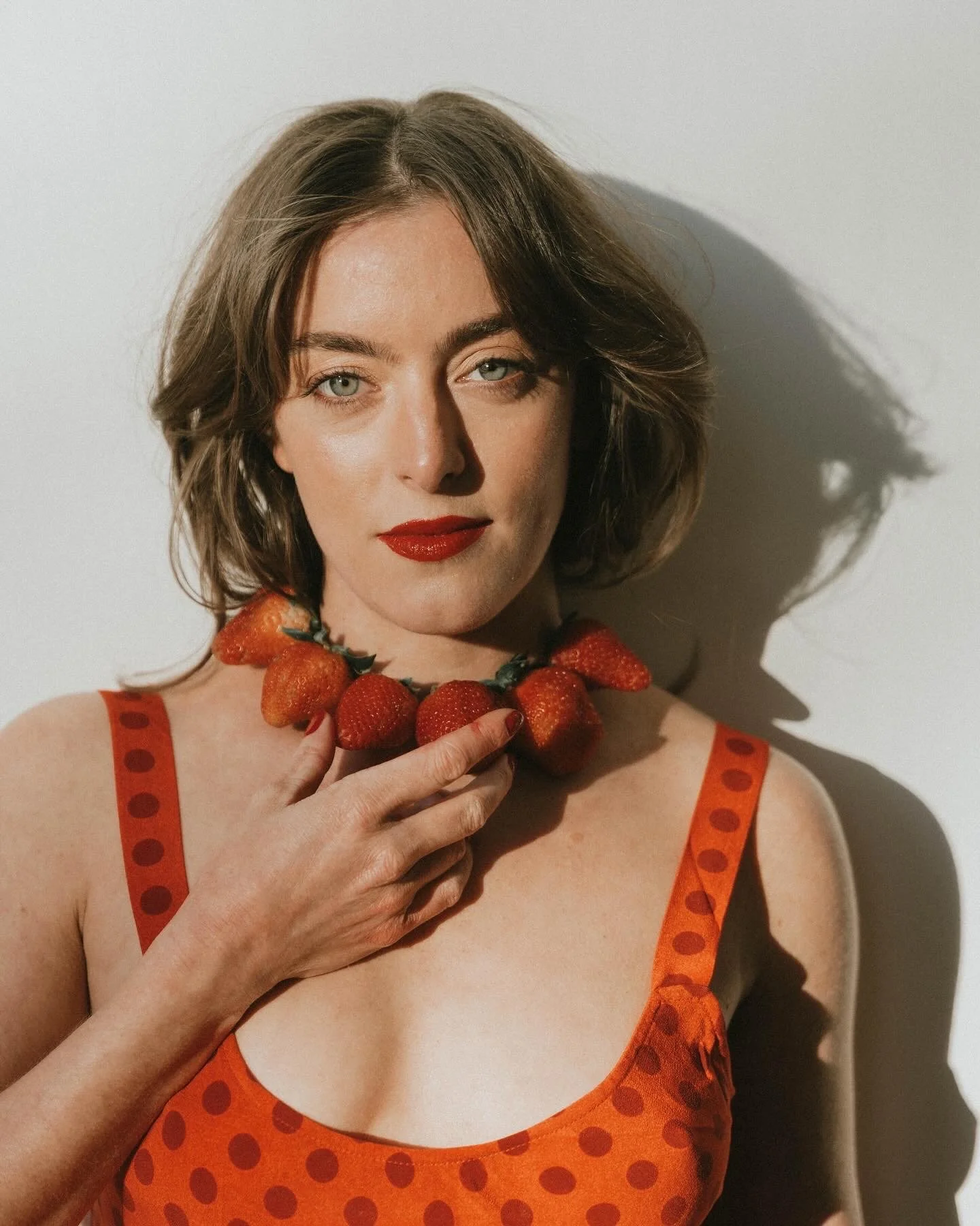 A woman with short light brown hair and blue eyes wears red lipstick and an orange dress with red polka dots. She holds a necklace made of strawberries close to her neck and looks at the camera. Creative Food Styling Photoshoot