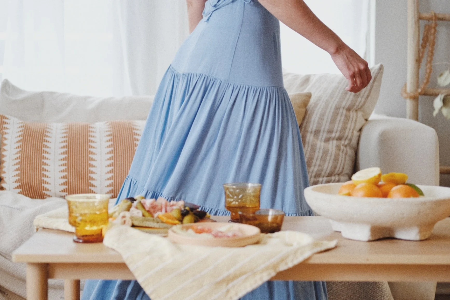 Person in a blue dress setting a table with food, including a bowl of oranges and glasses of juice, in a bright living room with a sofa and decorative pillows.