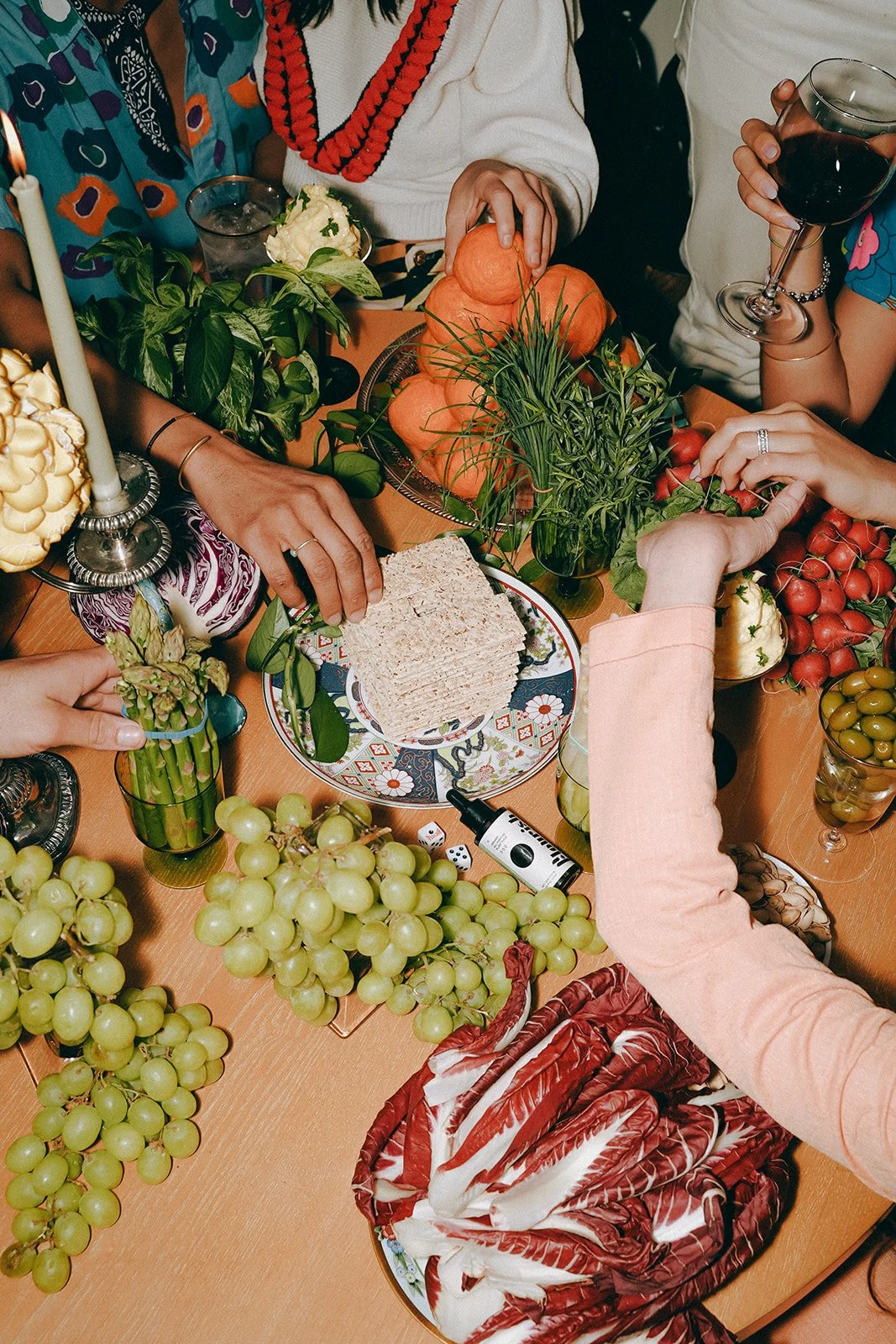 A table with various foods including grapes, cherry tomatoes, a layered cake, slices of bread, pumpkins, asparagus, and decorative candles, with people reaching for the food and holding a glass of red wine.