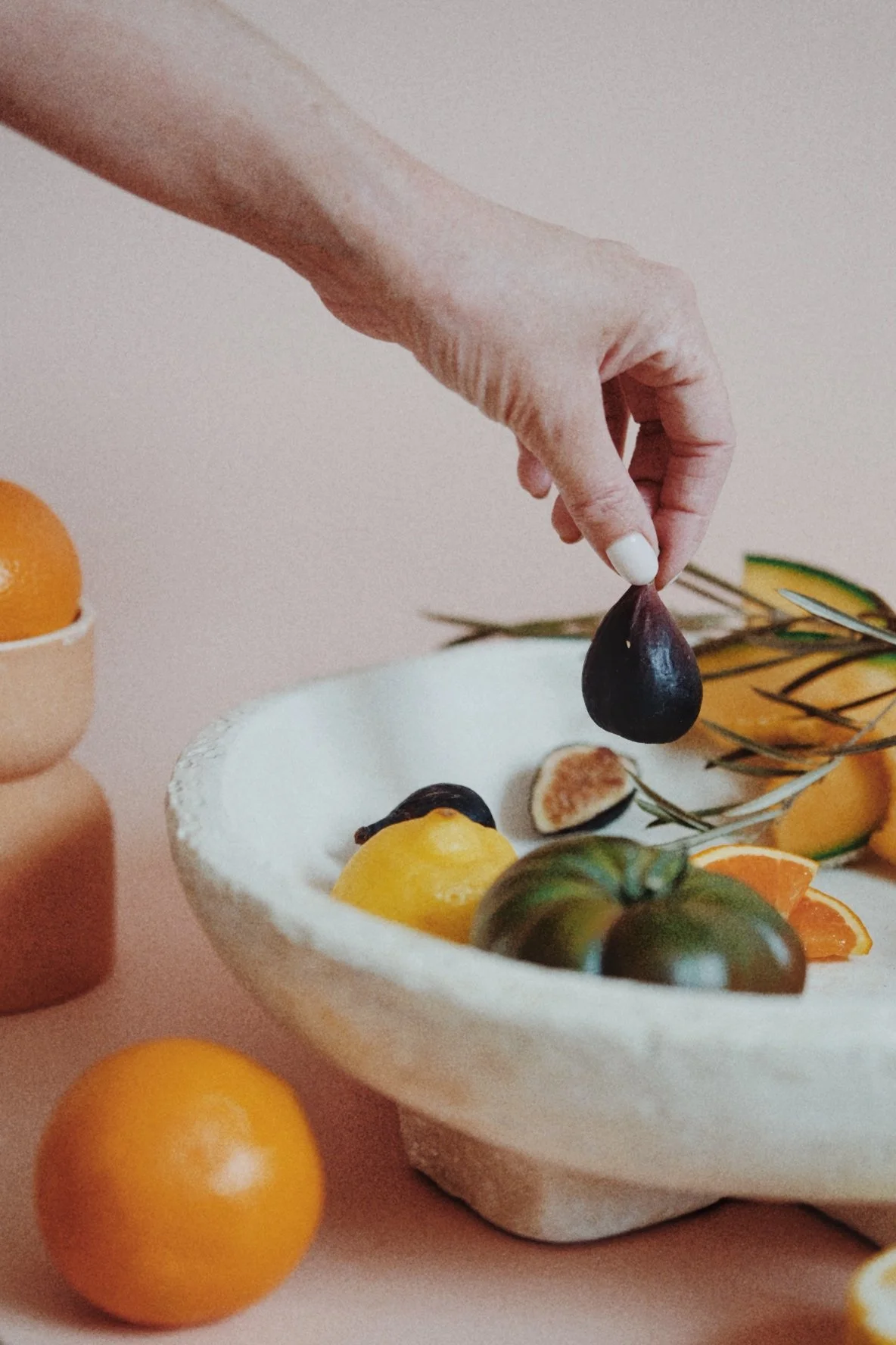 A human hand holding a dark purple fig over a white bowl filled with various fruits, including lemons, tomatoes, and an orange, with more fruits and greenery in the background.