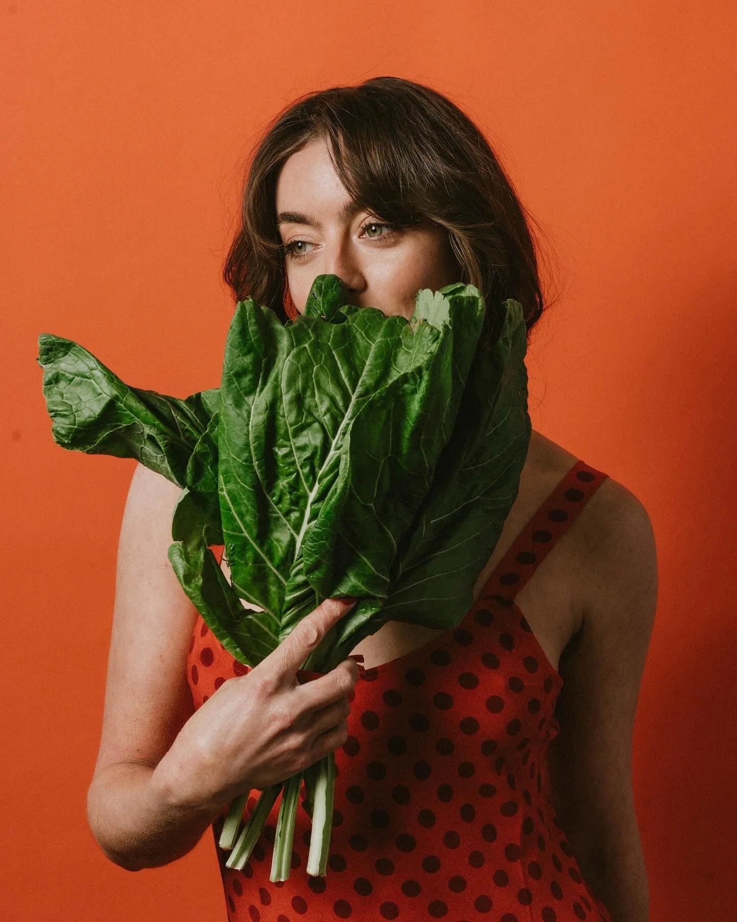 A woman with brown hair holds a bunch of large green leafy vegetables in front of her face, partially covering it, against an orange background. She wears a red dress with black polka dots.