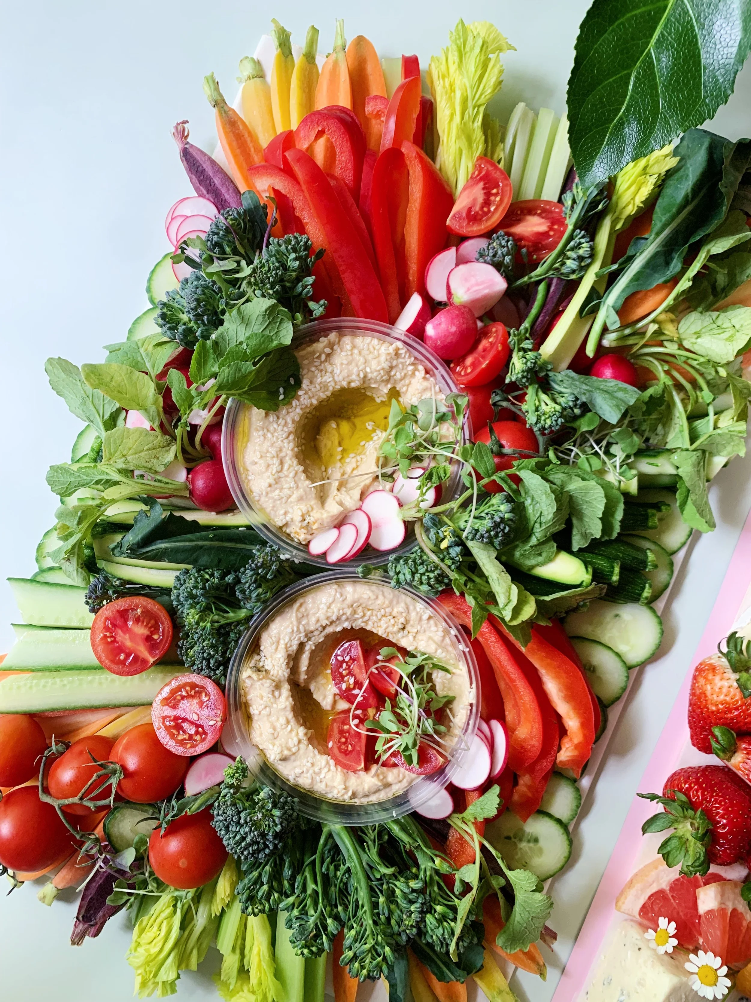A colorful display of fresh vegetables, including cherry tomatoes, radishes, cucumbers, carrots, bell peppers, celery, and leafy greens, arranged around two bowls of hummus garnished with herbs and vegetables.
