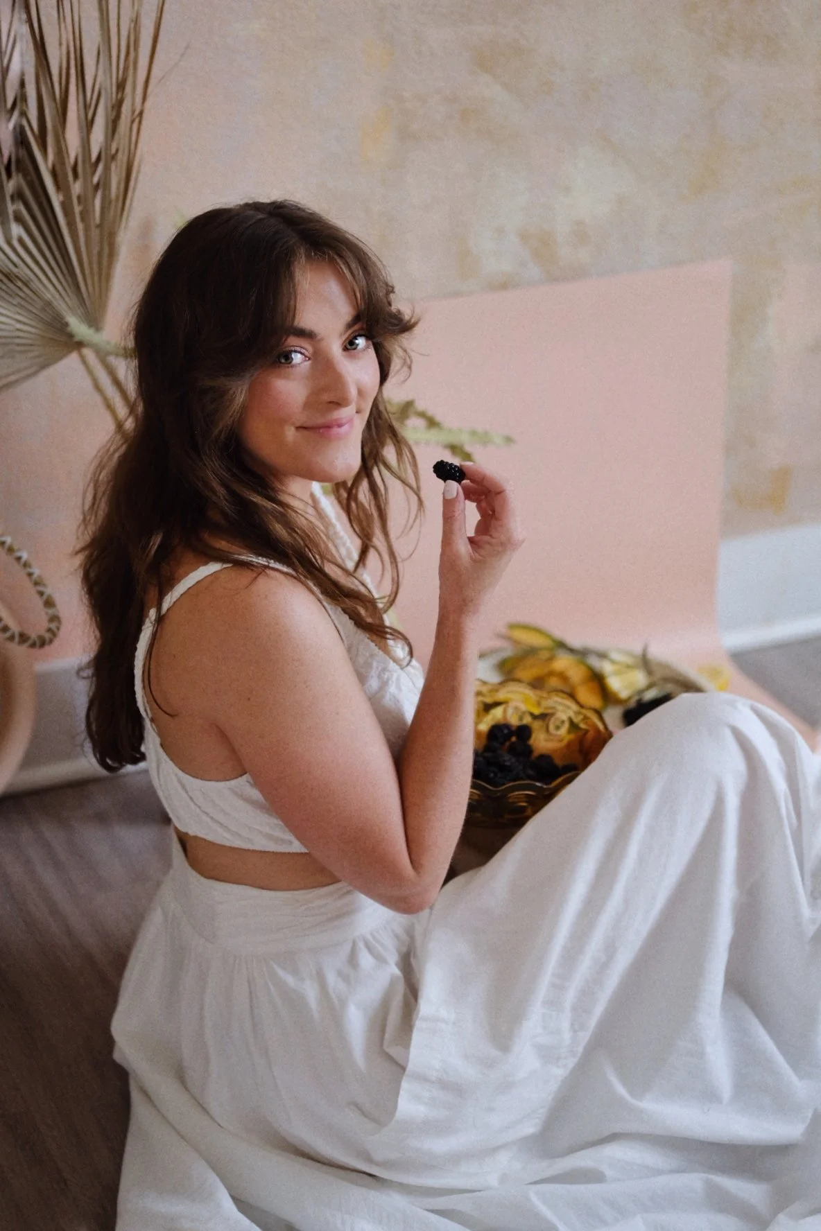 A woman with brown, wavy hair sitting on the floor, holding a blackberry, with a bowl of grapes and fruit in front of her, against a textured wall background.