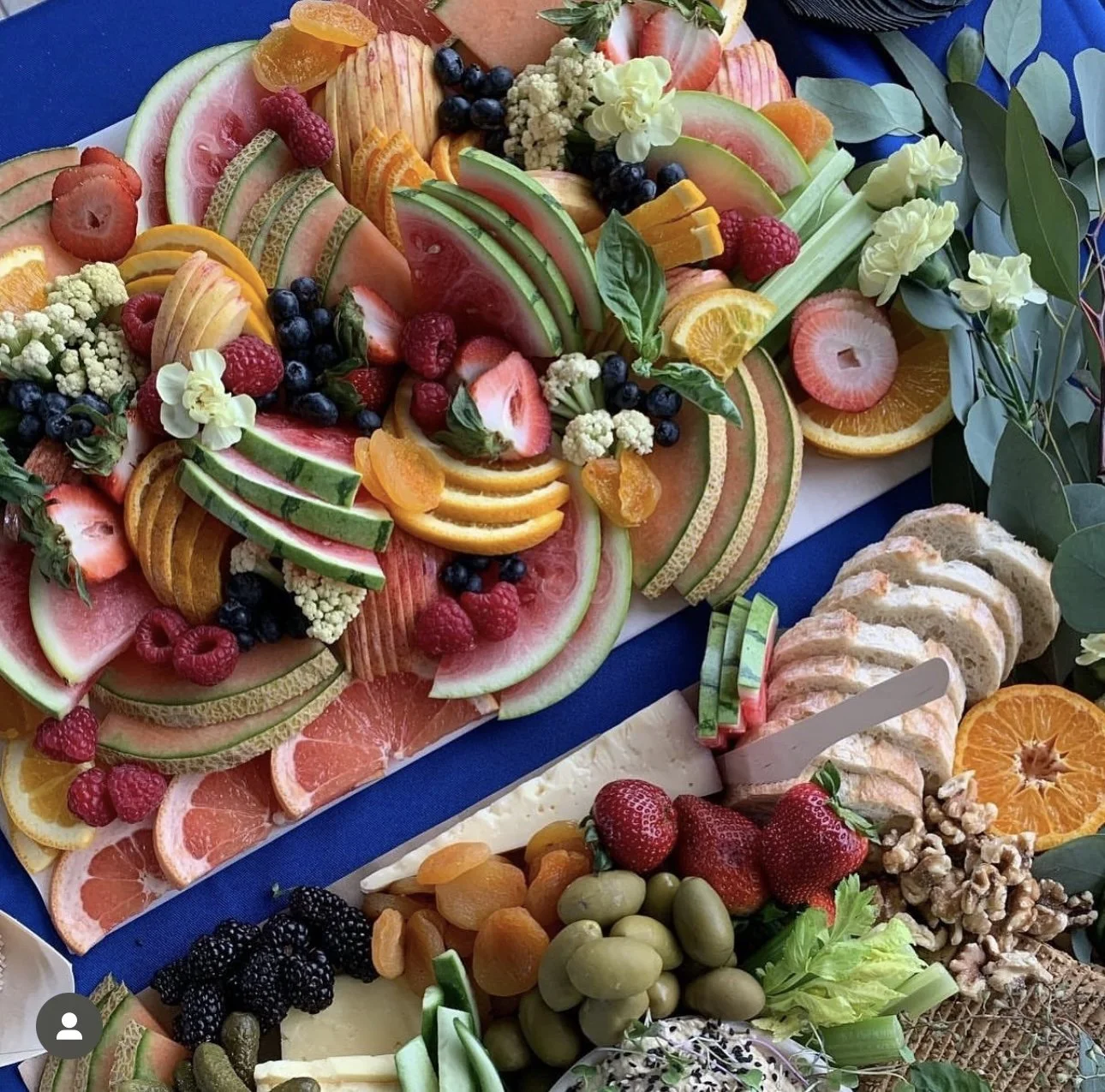 A large platter of assorted fresh fruits including watermelon, strawberries, raspberries, blueberries, blackberries, green grapes, black grapes, and orange slices, garnished with greenery and flowers. There are also sliced bread and cheese nearby.
