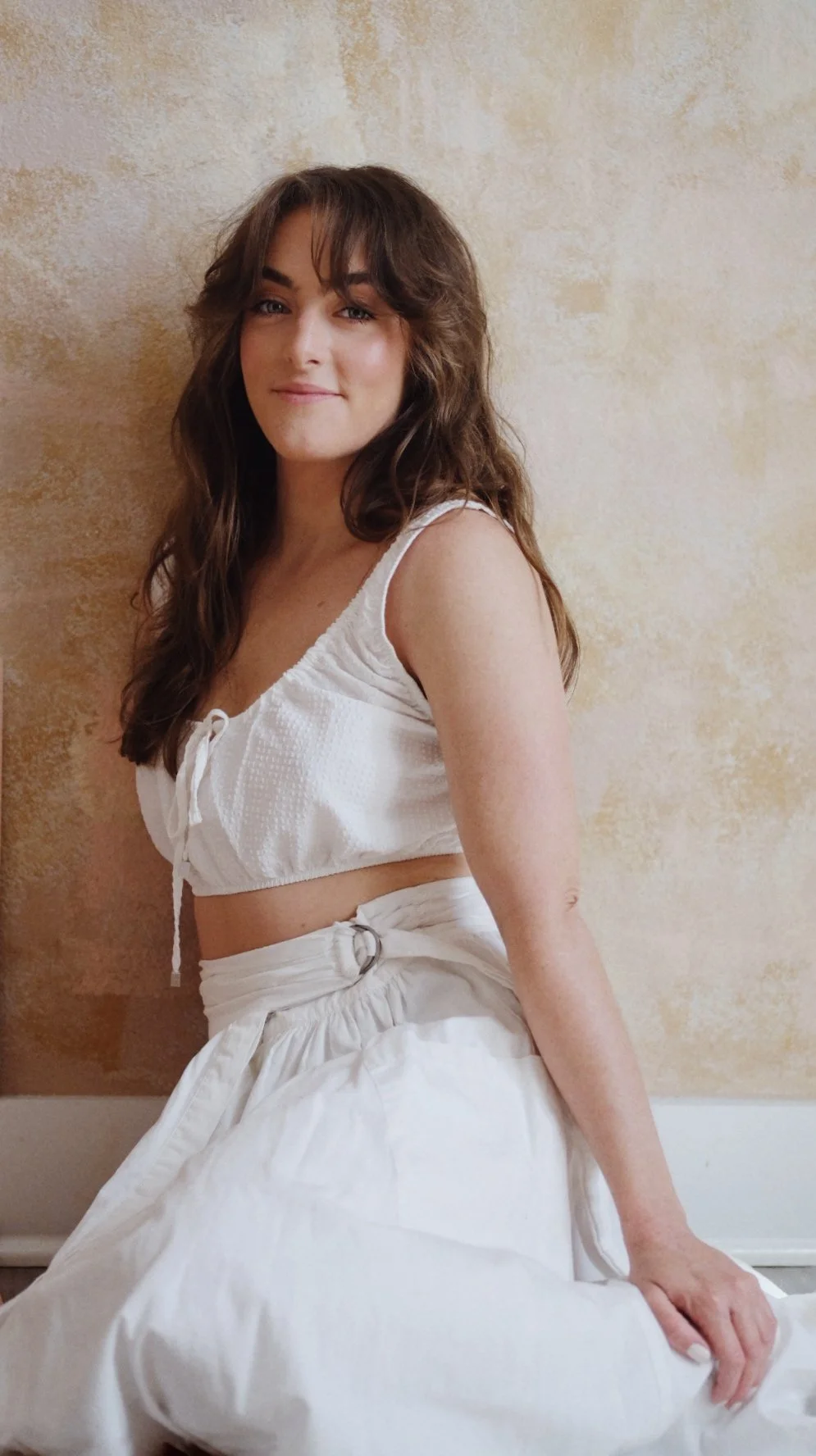 A young woman with long, curly brown hair and light skin, wearing a white sleeveless crop top and white high-waisted pants, sitting against a beige textured wall.