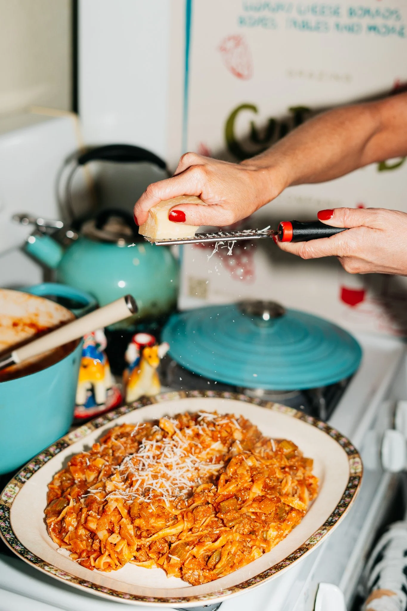 Person grating cheese over a plate of cooked pasta with sauce in a kitchen.