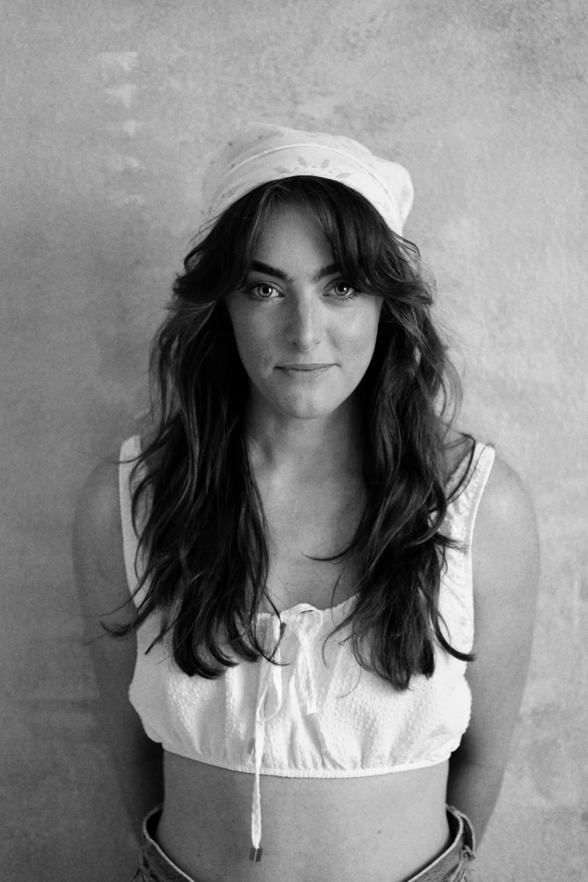 Black and white photo of a woman with long wavy hair, wearing a sleeveless top and a headscarf, looking directly at the camera.