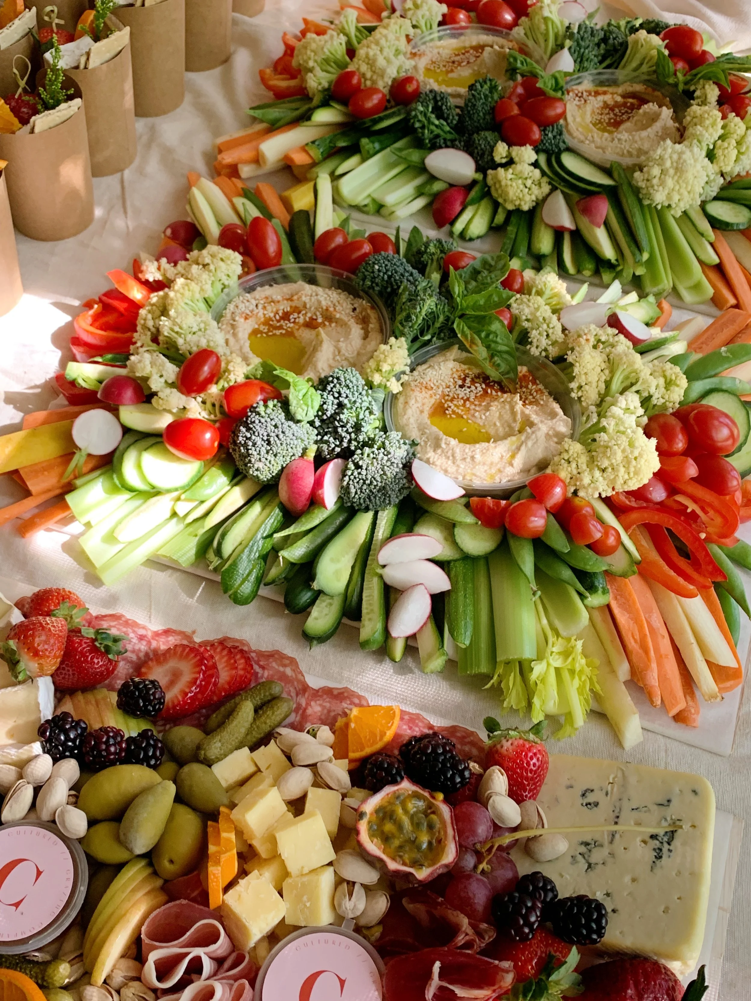 Colorful vegetable platter with cherry tomatoes, celery, carrots, radishes, broccoli, cauliflower, cucumber, and hummus, surrounded by a cheese and charcuterie board with strawberries, blackberries, blueberries, cheese cubes, nuts, cured meats, and a large wedge of blue cheese.