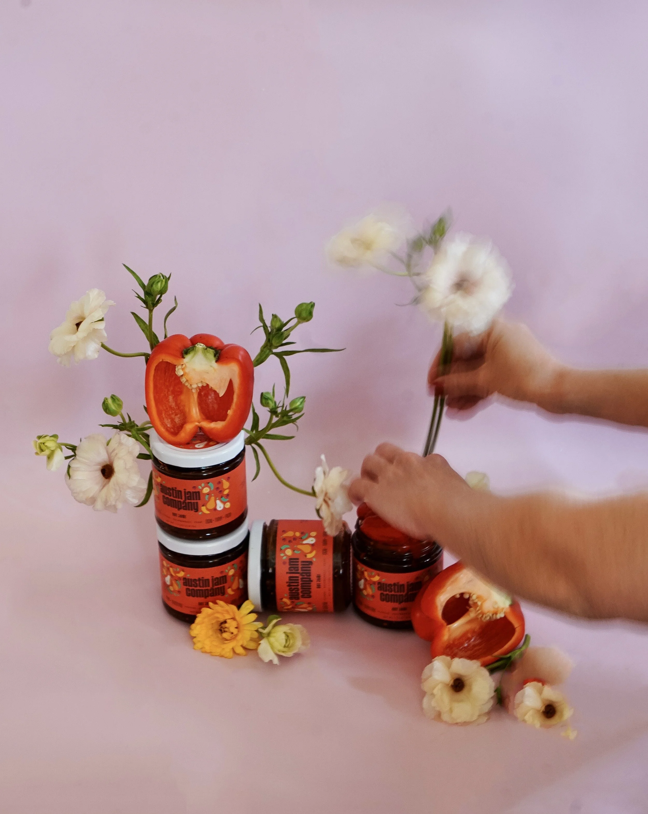 A person arranging flowers with jars of Austin Jam Company jam, with a cut red bell pepper and various flowers on a pink background.
