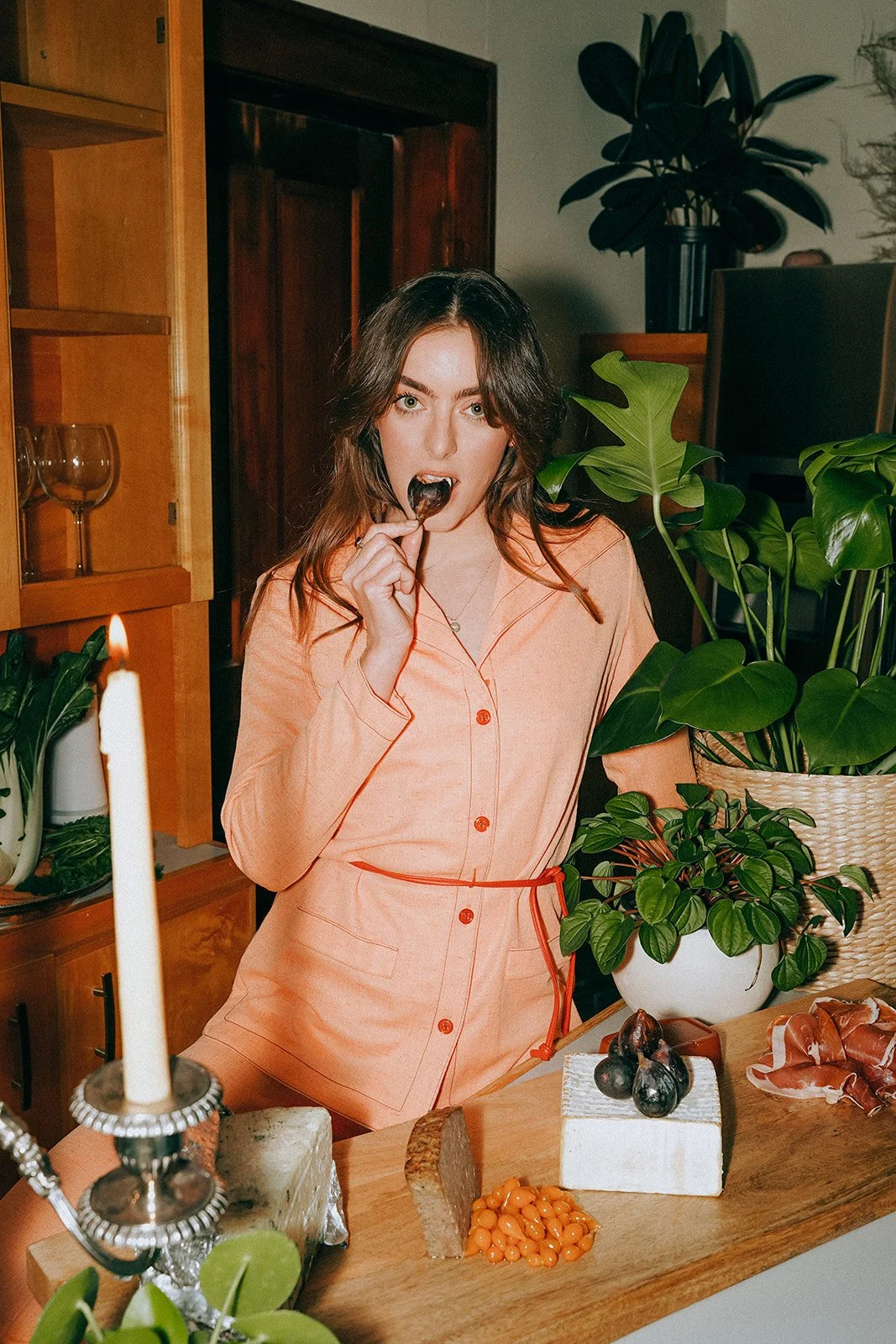 A woman wearing a peach-colored shirt is eating a black dish with a spoon in a kitchen or dining area decorated with green plants and cheese on a wooden table.