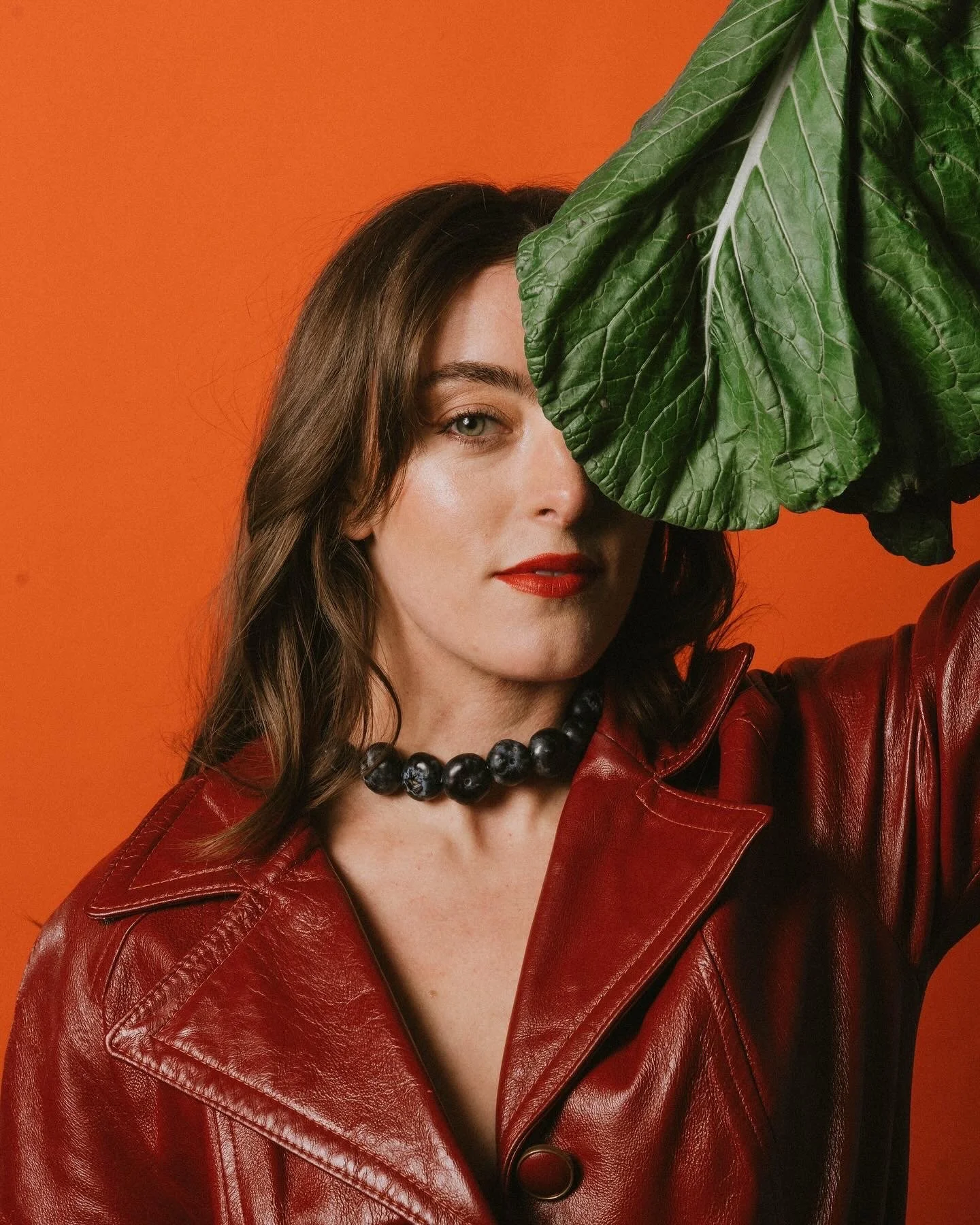 A woman with wavy brown hair, red lipstick, and green eyes, wearing a red leather jacket and a black beaded necklace, holding a large green leaf in front of her face against an orange background. Creative Food Styling Photoshoot. Cultured Creative