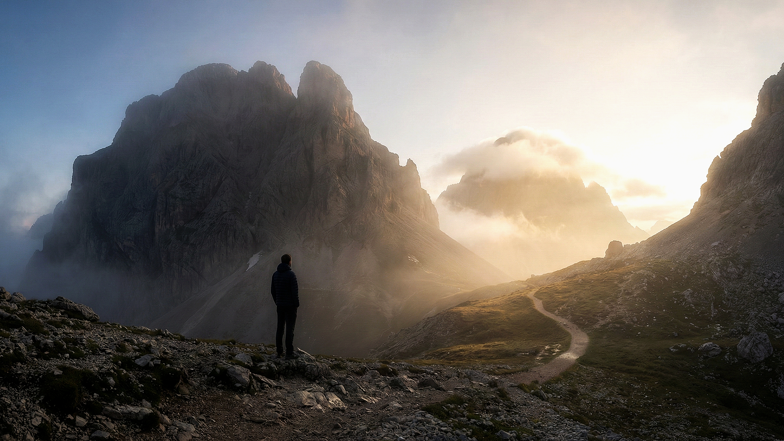 A person in a dark jacket standing on rocky terrain, looking at tall mountains in the distance with a winding trail leading toward them, under a partly cloudy sky with the sun shining through.