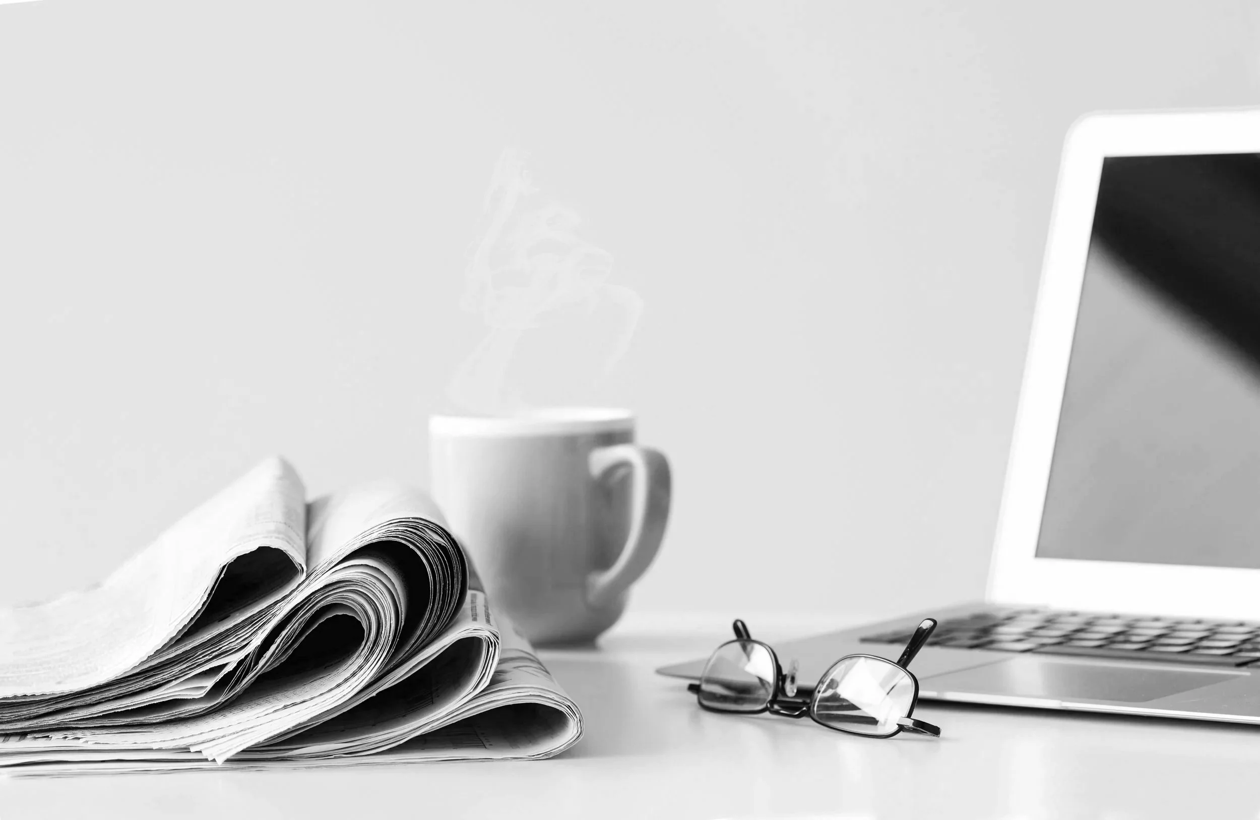 Desk with a laptop, folded newspapers, coffee mug, and glasses, representing reflection and thoughtful reading.