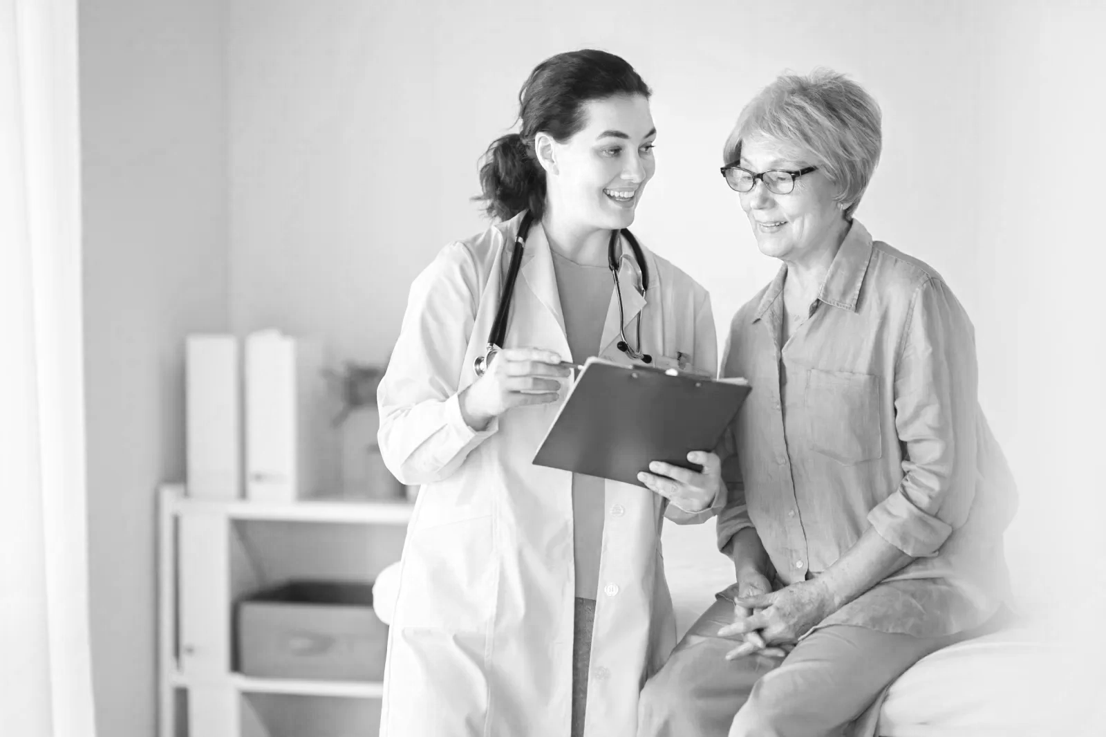 A female healthcare professional with a stethoscope around her neck shows a clipboard to an older female patient sitting on an examination table. The patient is smiling and wearing glasses.