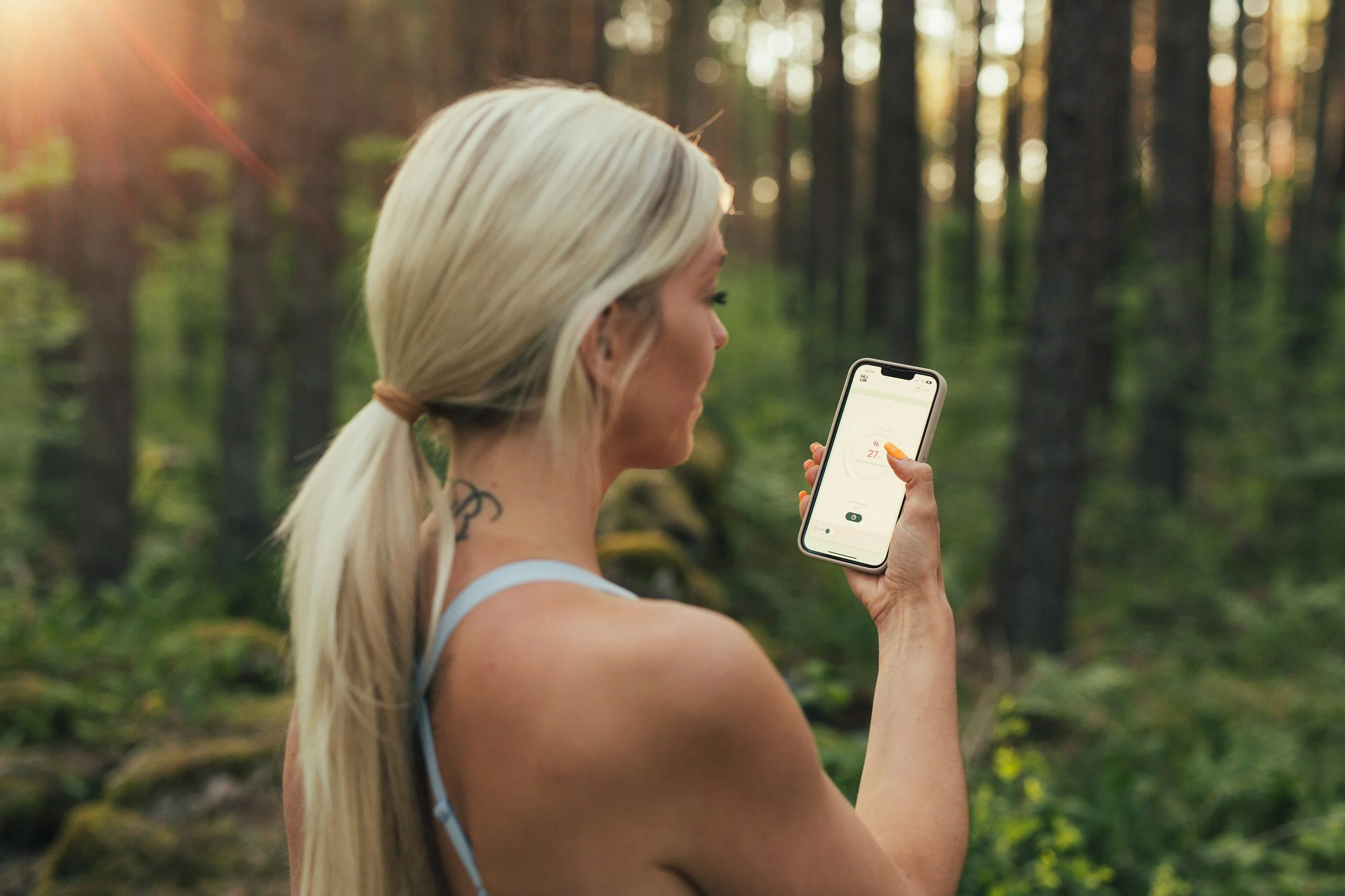 Woman with blonde hair in a ponytail using a smartphone in a wooded forest during sunset.