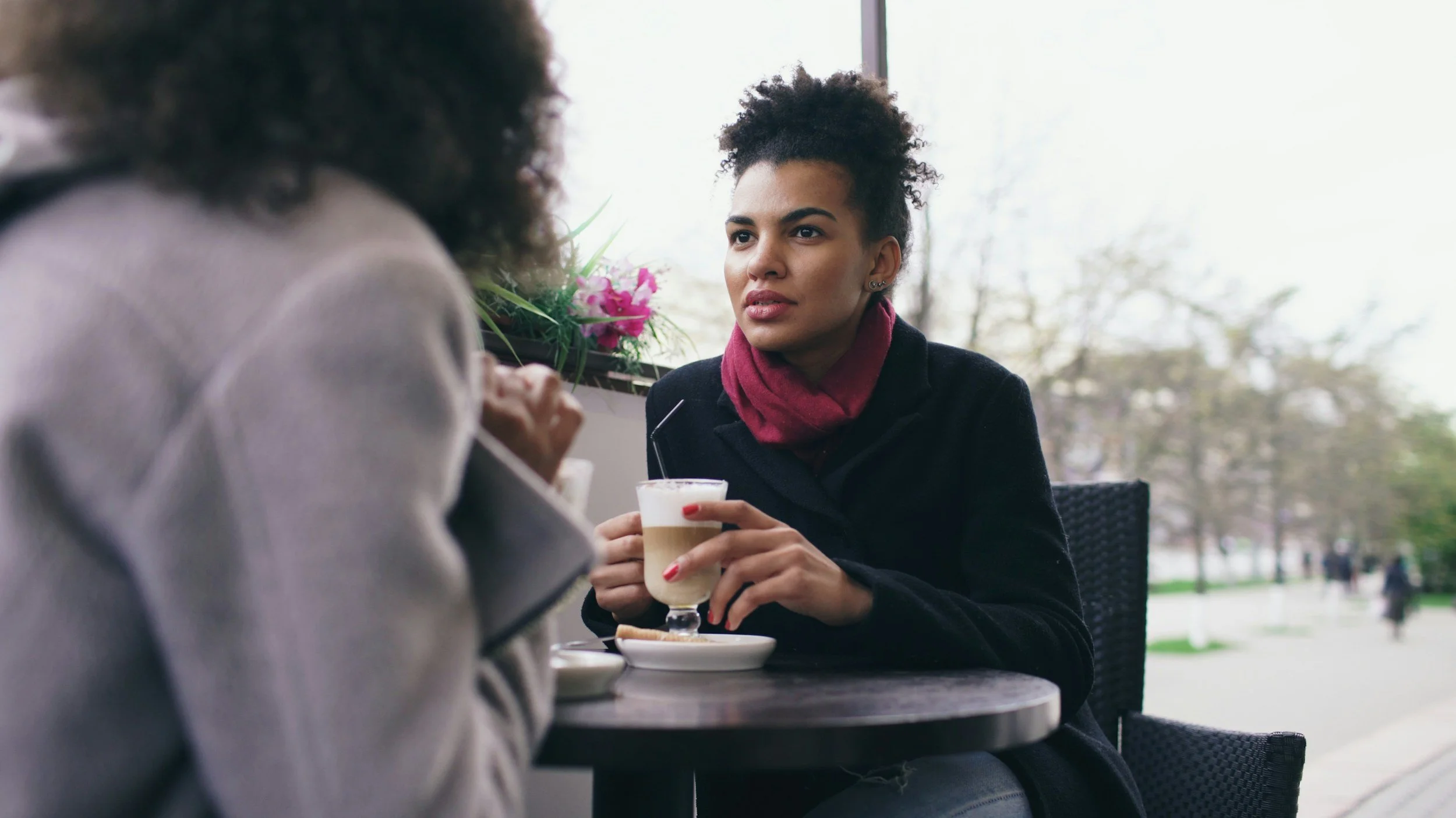 Two women sitting at a table in a cafe, one is visible and the other has her back to the camera. The woman facing the camera has short curly hair, is wearing a black coat and a red scarf, and is holding a glass of coffee or latte. There is a flowerpot with pink flowers on the window behind her, and a window showing an outdoor scene with trees and people walking.