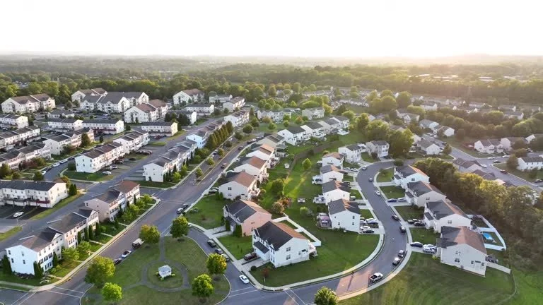 Aerial view of a suburban neighborhood with rows of white houses, green yards, and parking lots, surrounded by trees at sunset.