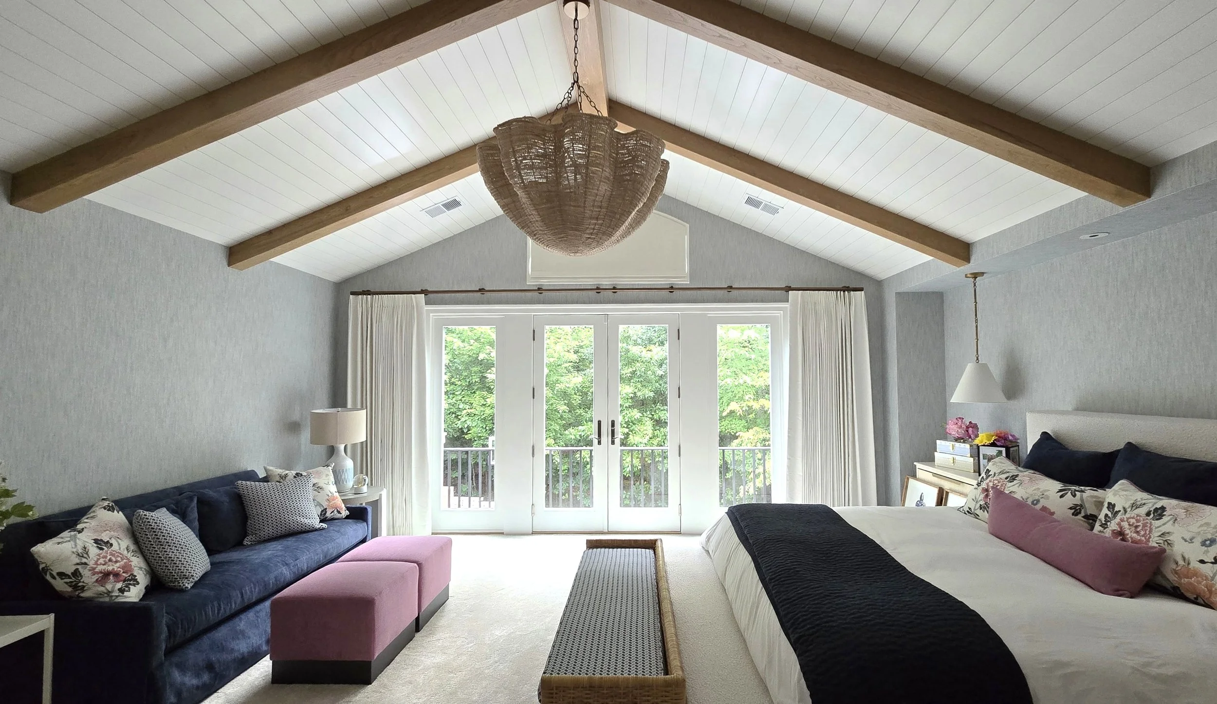 A bedroom with a vaulted ceiling featuring exposed wooden beams, a large bed with black, pink, and floral pillows, a white nightstand with a lamp, a navy blue sofa with floral and striped pillows, a pink ottoman, and sliding glass doors leading to a balcony with green trees outside.