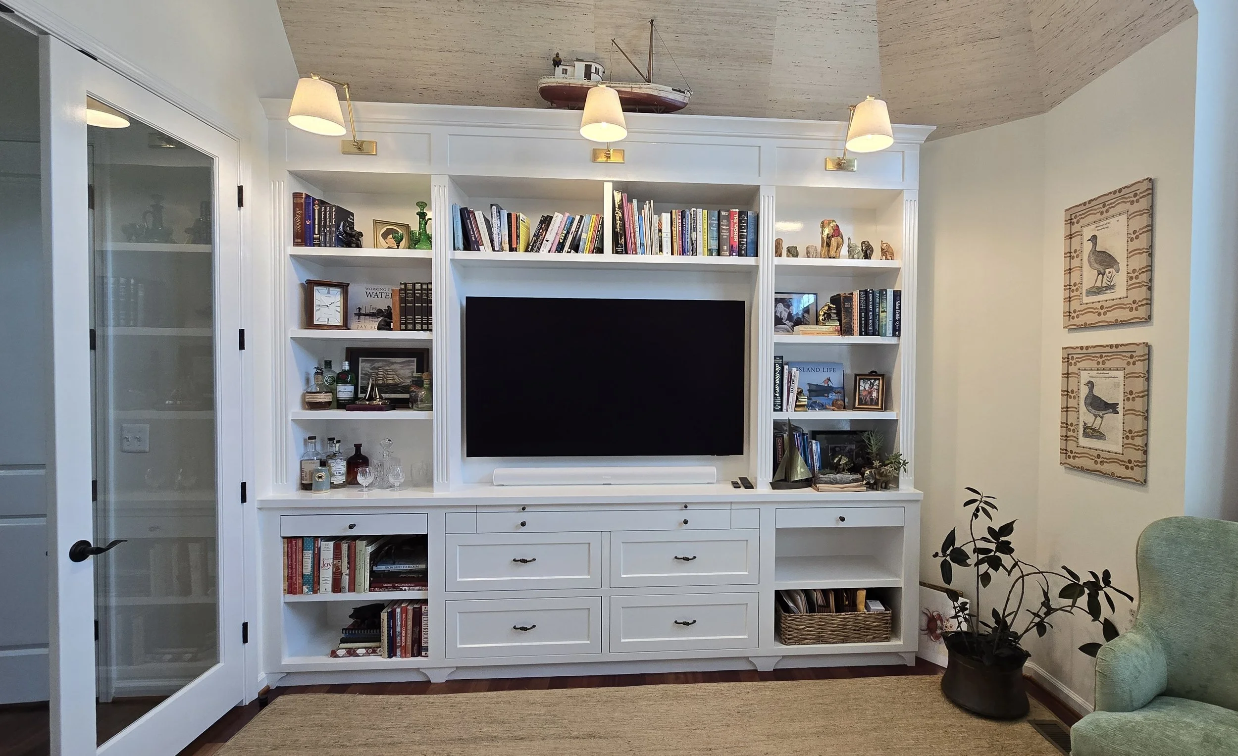 Living room entertainment center with white shelves filled with books, decorative items, and small collectibles. A large flat-screen TV is centered on the unit. There is a soundbar below the TV. To the right of the unit, a green armchair is visible, with a potted plant nearby. On the wall to the right, two framed pictures of ducks are hung. The room has a beige carpet and a wooden floor.