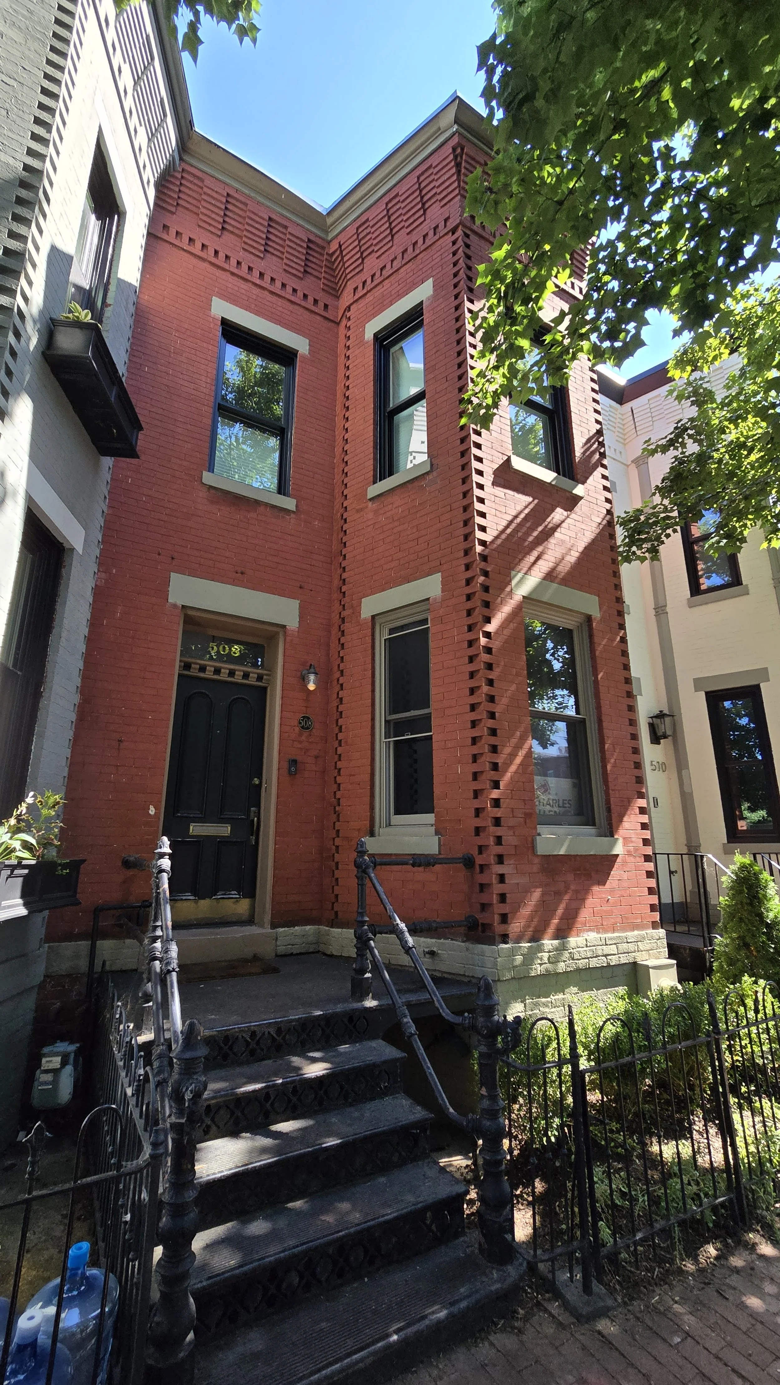 A brick townhouse before painting A three-story red brick townhouse with black front door, surrounded by black iron railing, with trees casting shadows on the facade in sunny weather.