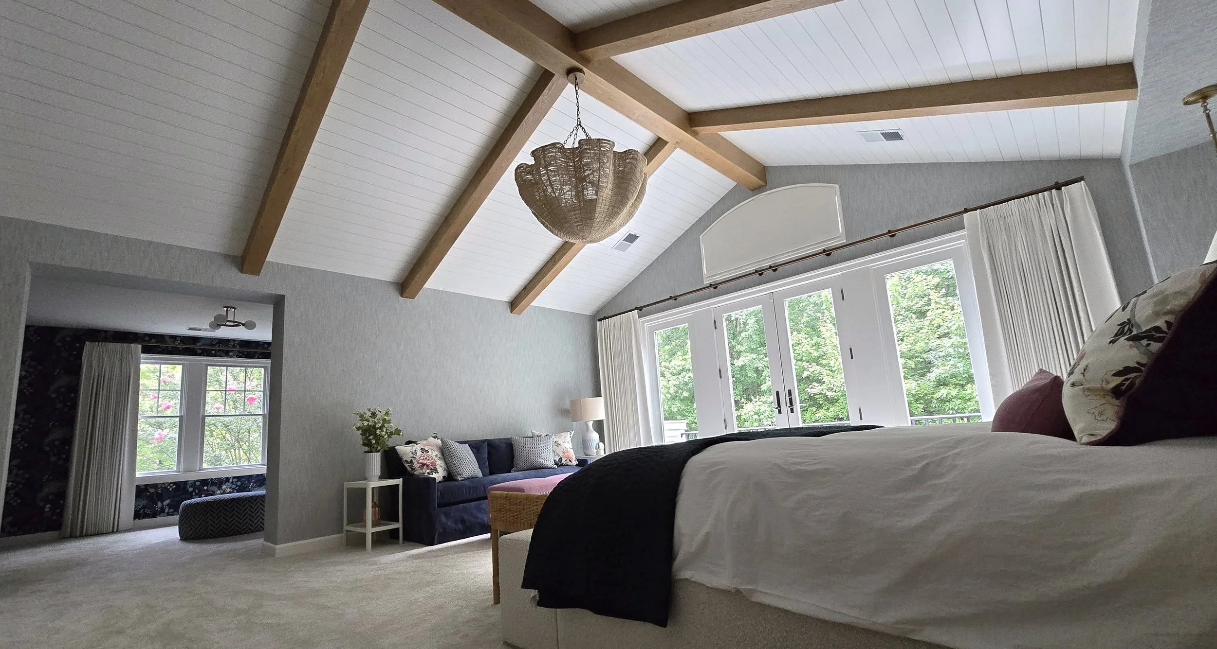 Bedroom with vaulted ceiling, wooden beams, large window with white curtains, bed with white bedding and decorative pillows, dark sofas with floral and patterned pillows, small side table with a lamp, and a view of lush green trees outside.