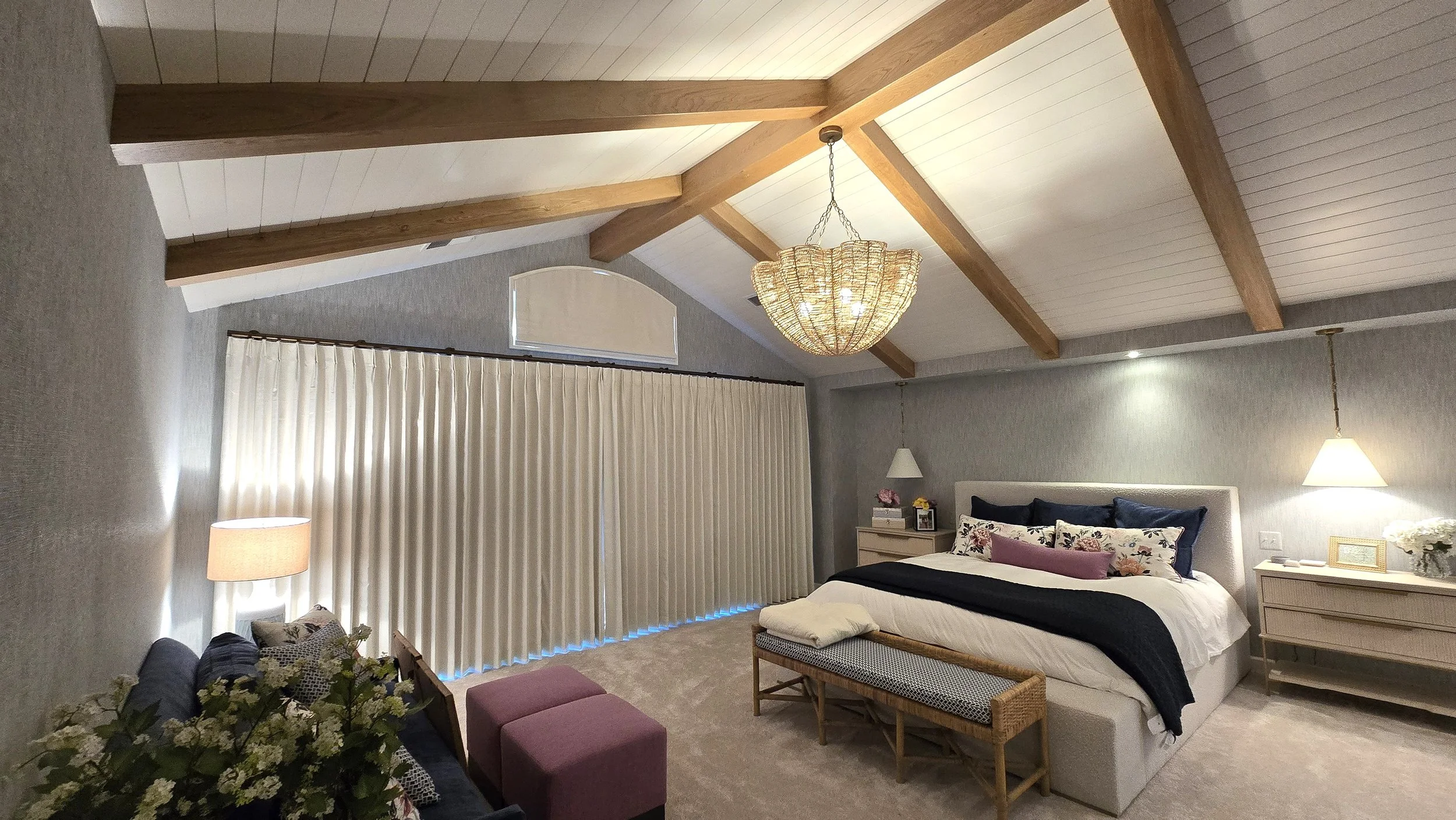 A spacious bedroom with a high, vaulted ceiling featuring exposed wooden beams and a chandelier. The room has large vertical blinds covering a window, a bed with multiple pillows, bedside tables with lamps, a floral arrangement on one side, and a small bench at the foot of the bed.