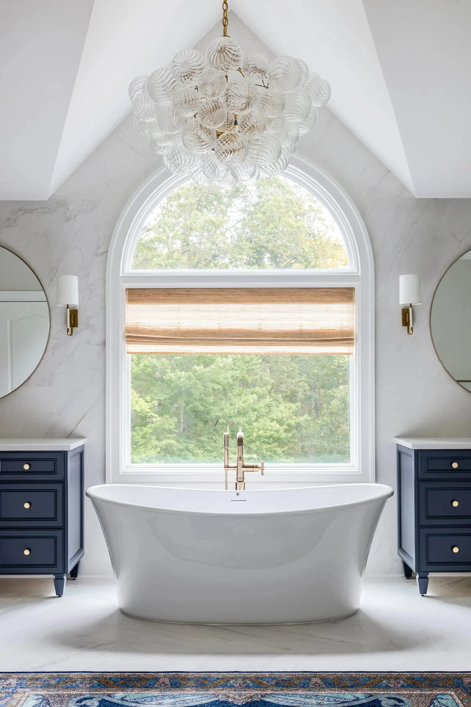 A luxurious bathroom features a white freestanding bathtub positioned in front of a large arched window with a view of green trees outside. Two navy blue vanities with gold accents are on either side of the tub, each topped with a mirror and a wall sconce. A modern chandelier with glass orbs hangs from the ceiling, adding elegance to the space.