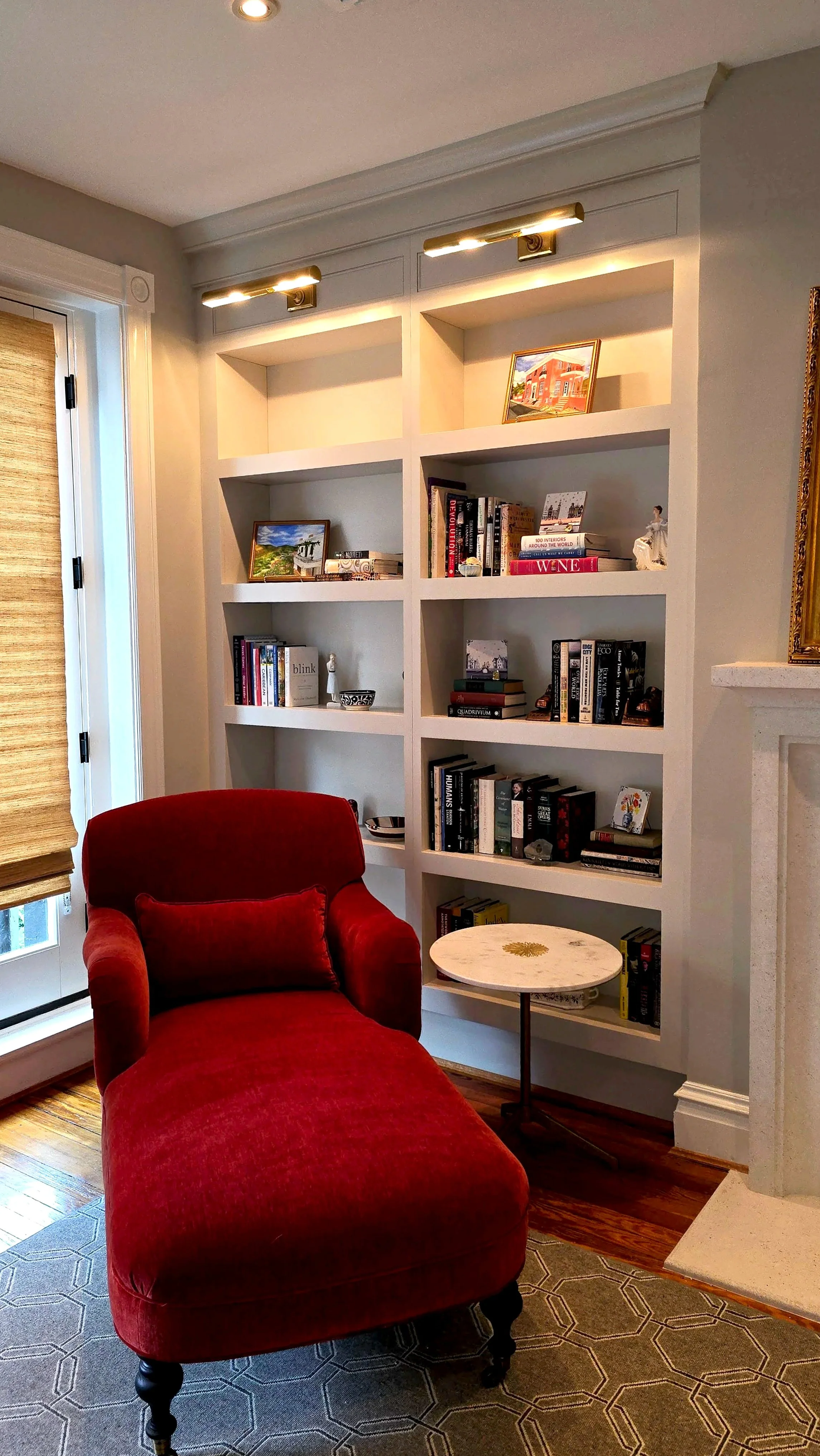 Living room corner with red chaise lounge, small round white side table, built-in white bookshelf filled with books and decorative items, window with woven shade, and hardwood floor.