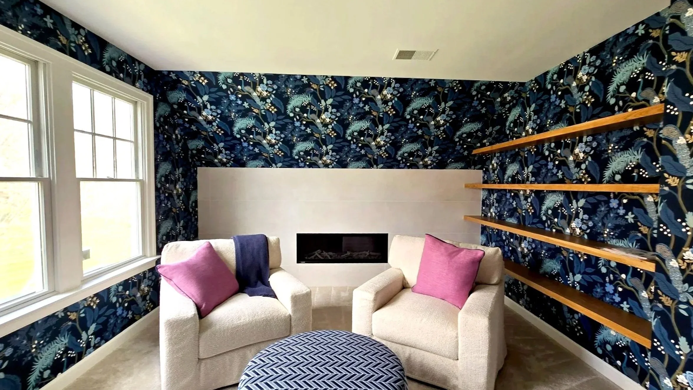Living room with two beige armchairs, pink and purple pillows, a modern fireplace, a navy floral accent wall, large windows, and wooden shelves.