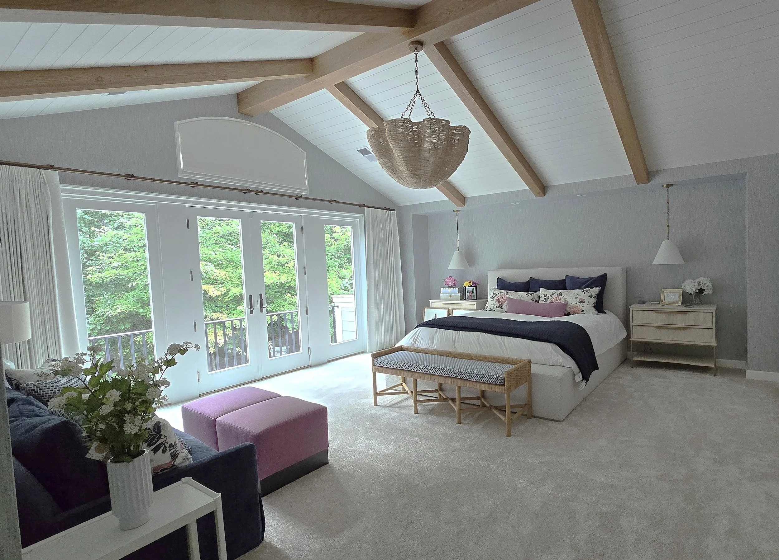 A bright bedroom with a vaulted ceiling, exposed wood beams, and a chandelier. There's a large bed with multiple pillows on the right, nightstands on each side, and large glass doors leading to a balcony with green trees outside. A sofa with a pink ottoman is on the left side.