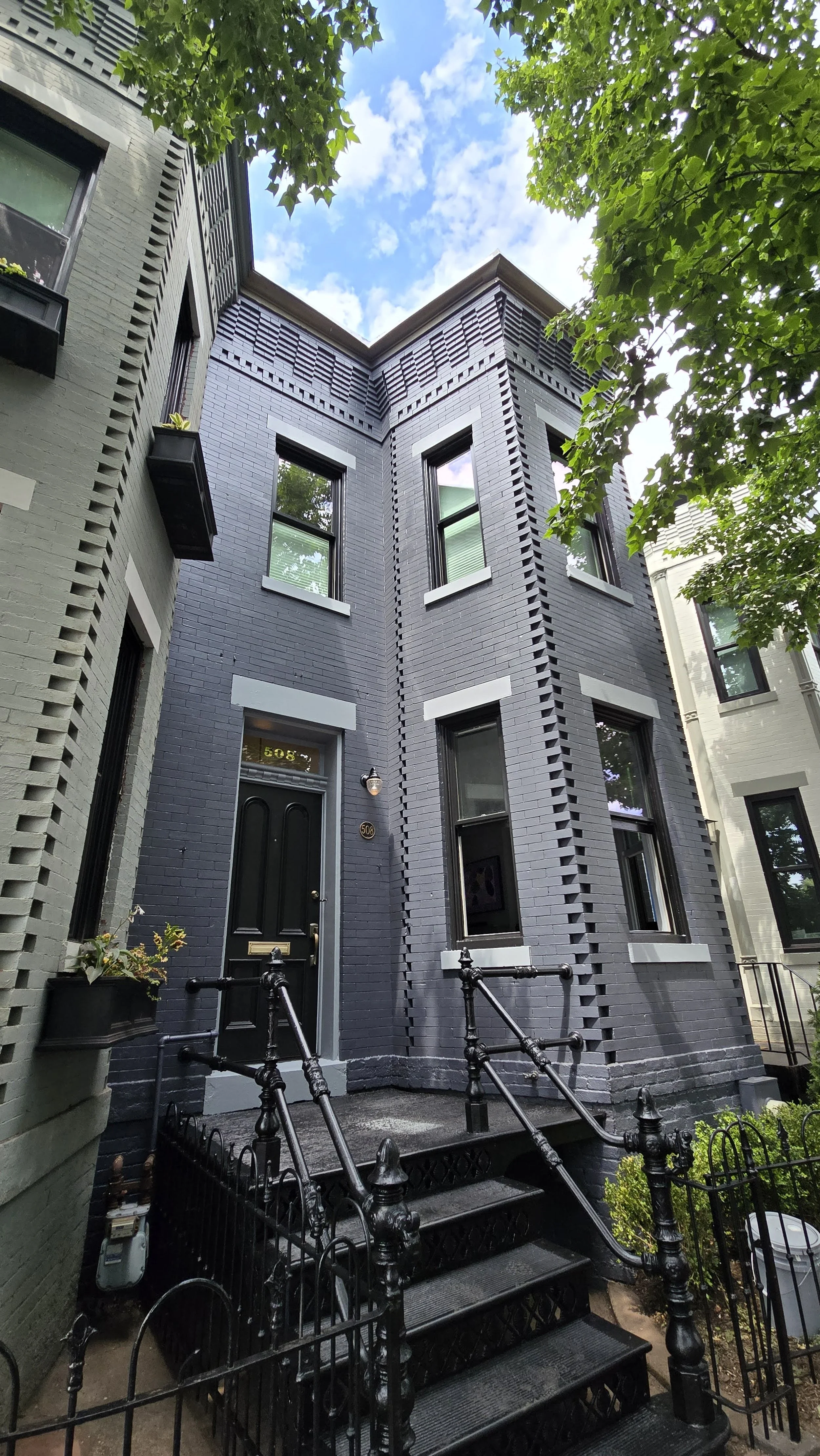 A brick townhouse we recently painted in washington DC with a front entrance accessed by black metal stairs and railings, surrounded by a small fenced yard, with trees and blue sky above.