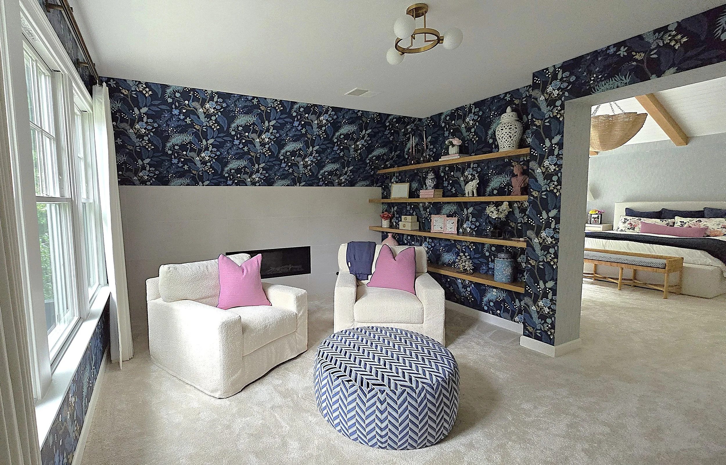 Living room with a beige armchair and loveseat, a blue and white patterned pouf, a salt and pepper corner fireplace, large windows, navy floral wallpaper accent wall, and shelves with decorative items. Bedroom visible in the background.