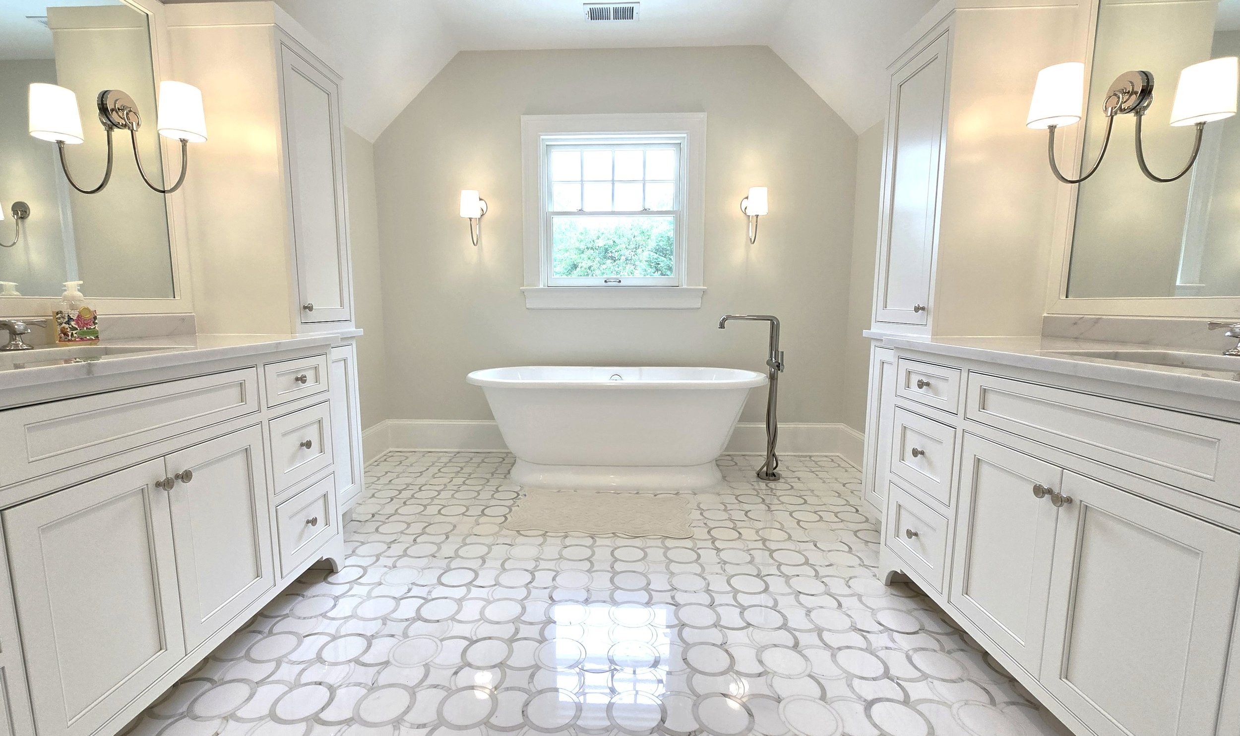 Bright bathroom with white cabinetry, a freestanding bathtub by a window, and wall-mounted light fixtures.