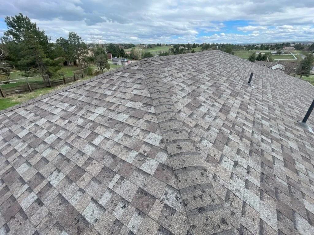 A rooftop covered with multiple rows of brown and gray shingles, with a central ridge and several vents, overlooking a rural landscape with trees, fences, and fields under a cloudy sky.